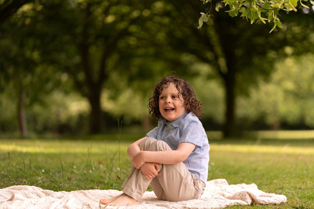 Happy curly-haired child sitting cross-legged on a blanket in a lush green park, smiling joyfully.