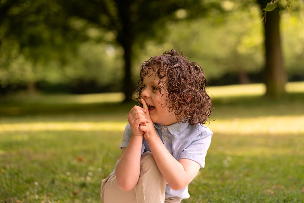 Curly-haired child clasping hands near mouth in a sunny green park, appearing surprised or excited.