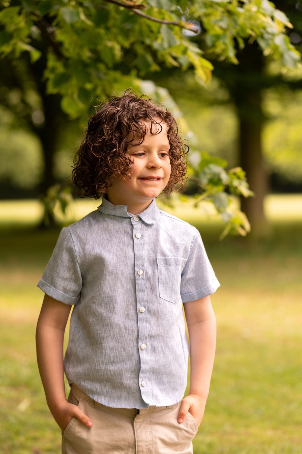 Curly-haired boy in a light blue linen shirt smiling outdoors in a sunny green park.