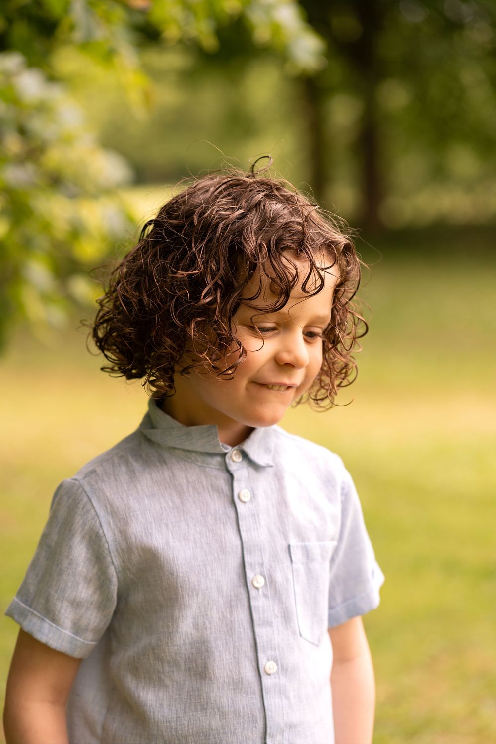 A smiling curly-haired boy in a grey short-sleeve shirt stands outdoors in a sunny green park, looking downward.