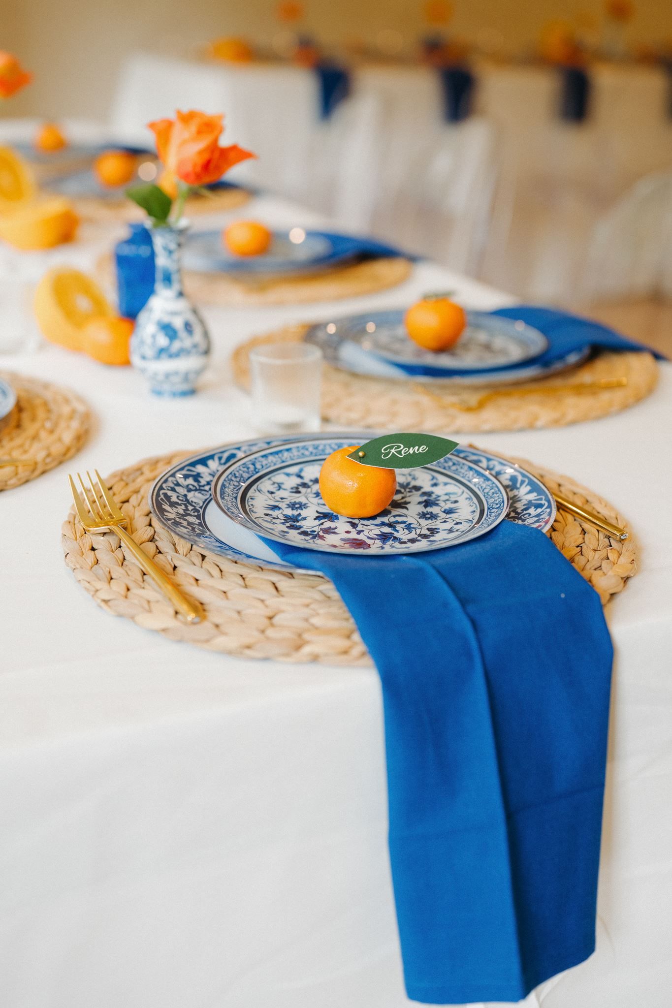 Blue and white porcelain plates with gold cutlery, blue napkins, and orange citrus fruits on woven placemats at an elegant table.