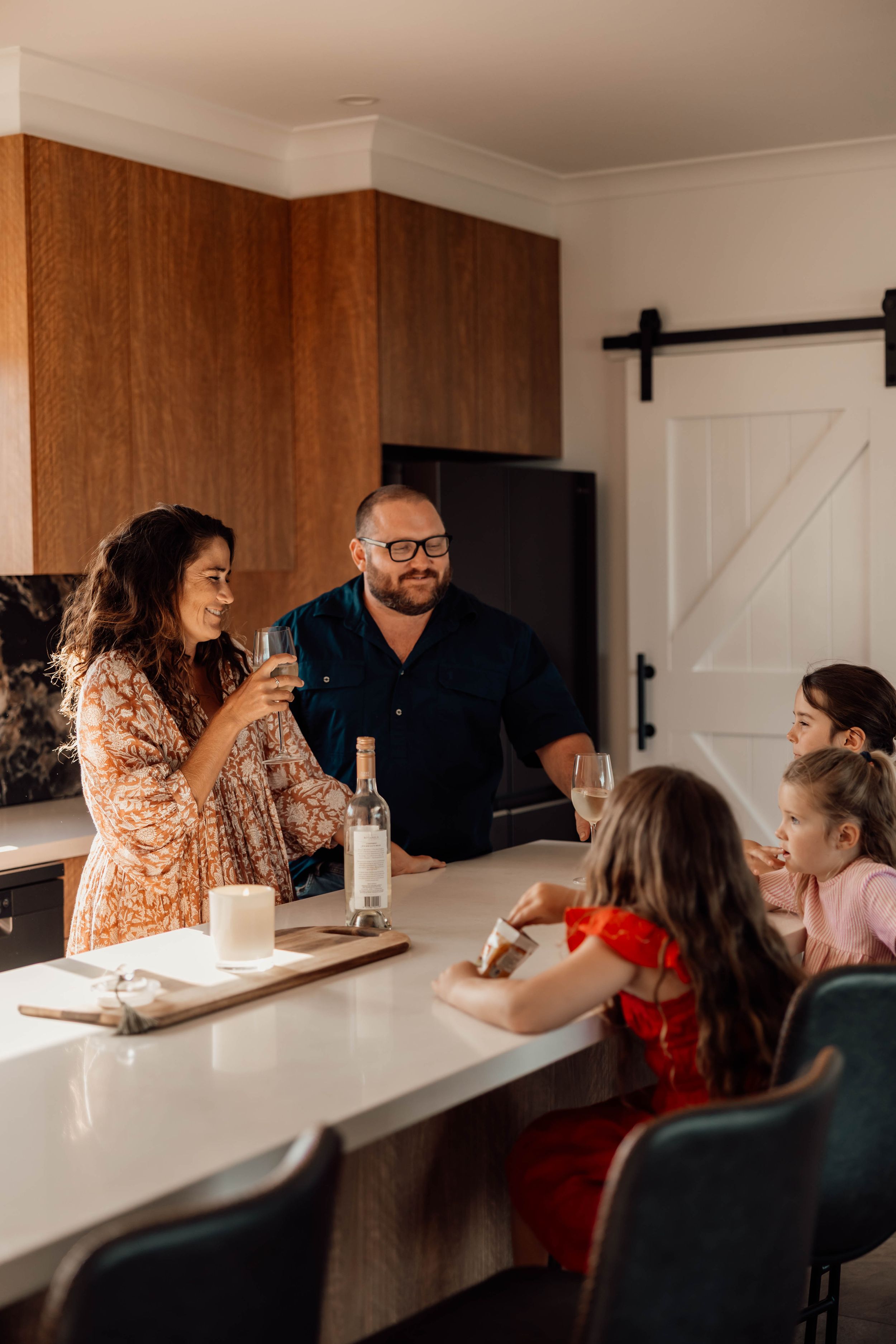 Family gathered around a white kitchen island, parents toasting wine glasses while children sit nearby in a modern home.