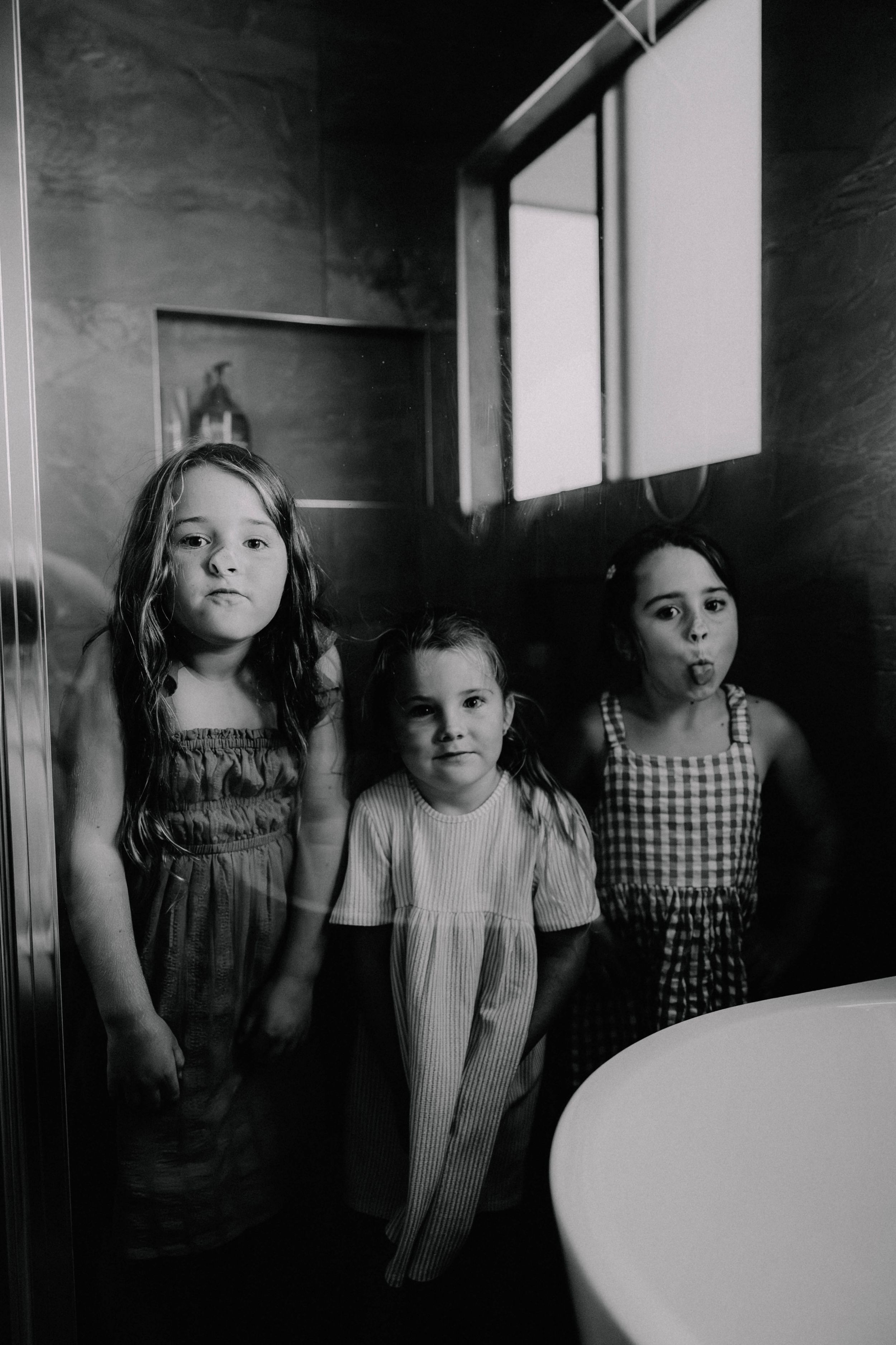 Three young girls stand together in a bathroom, captured in black and white photography.
