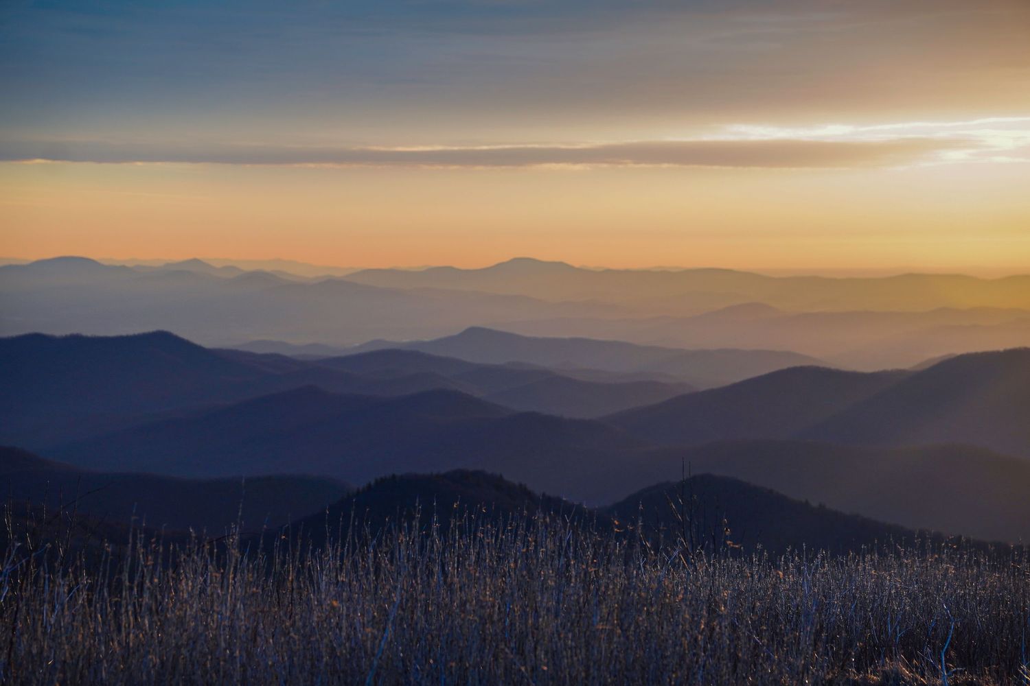 Golden sunset light illuminates layered mountain ridges with frost-covered grasses in the foreground.