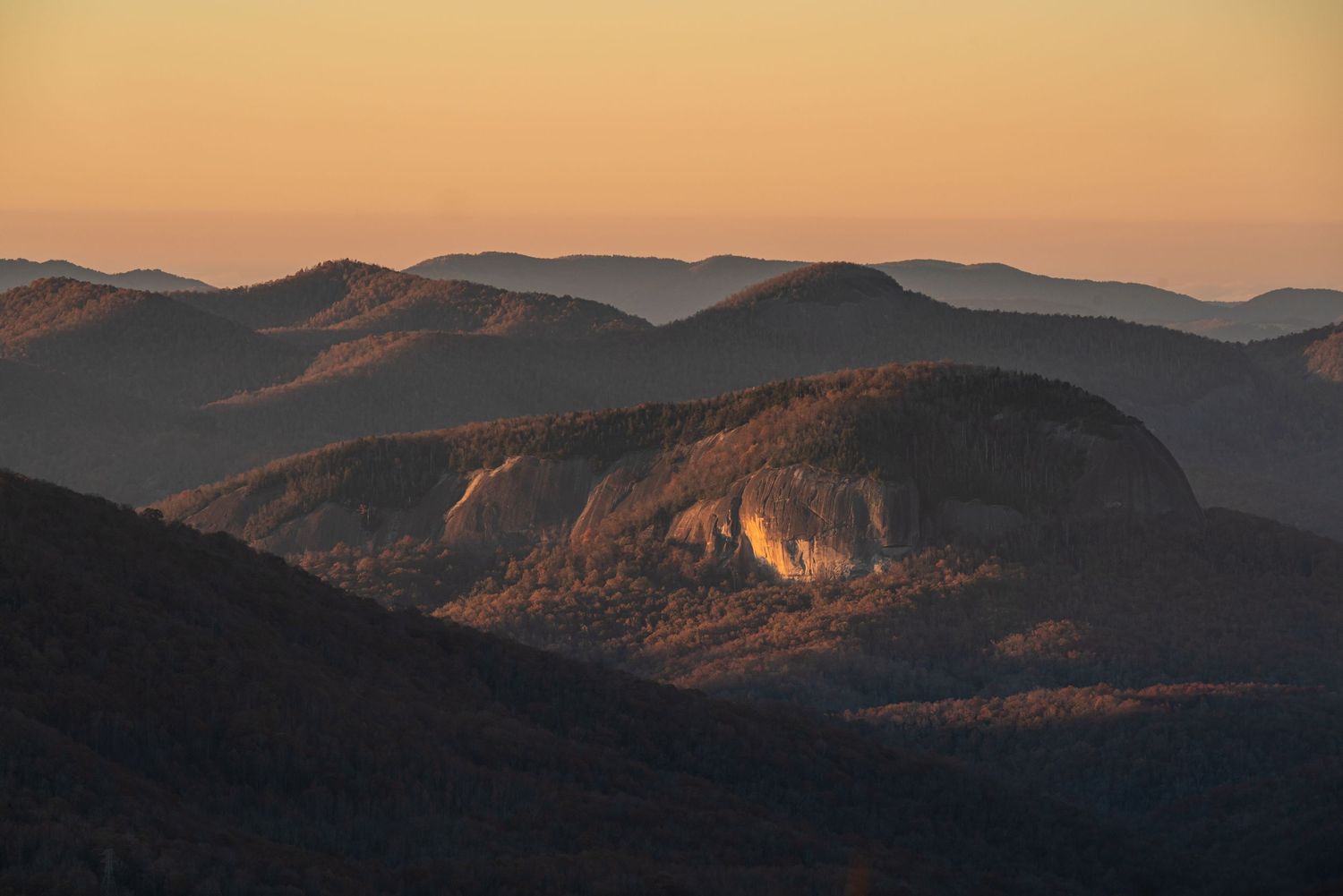 A rocky mountain summit catches warm sunset light surrounded by rolling forested ridgelines at dusk.