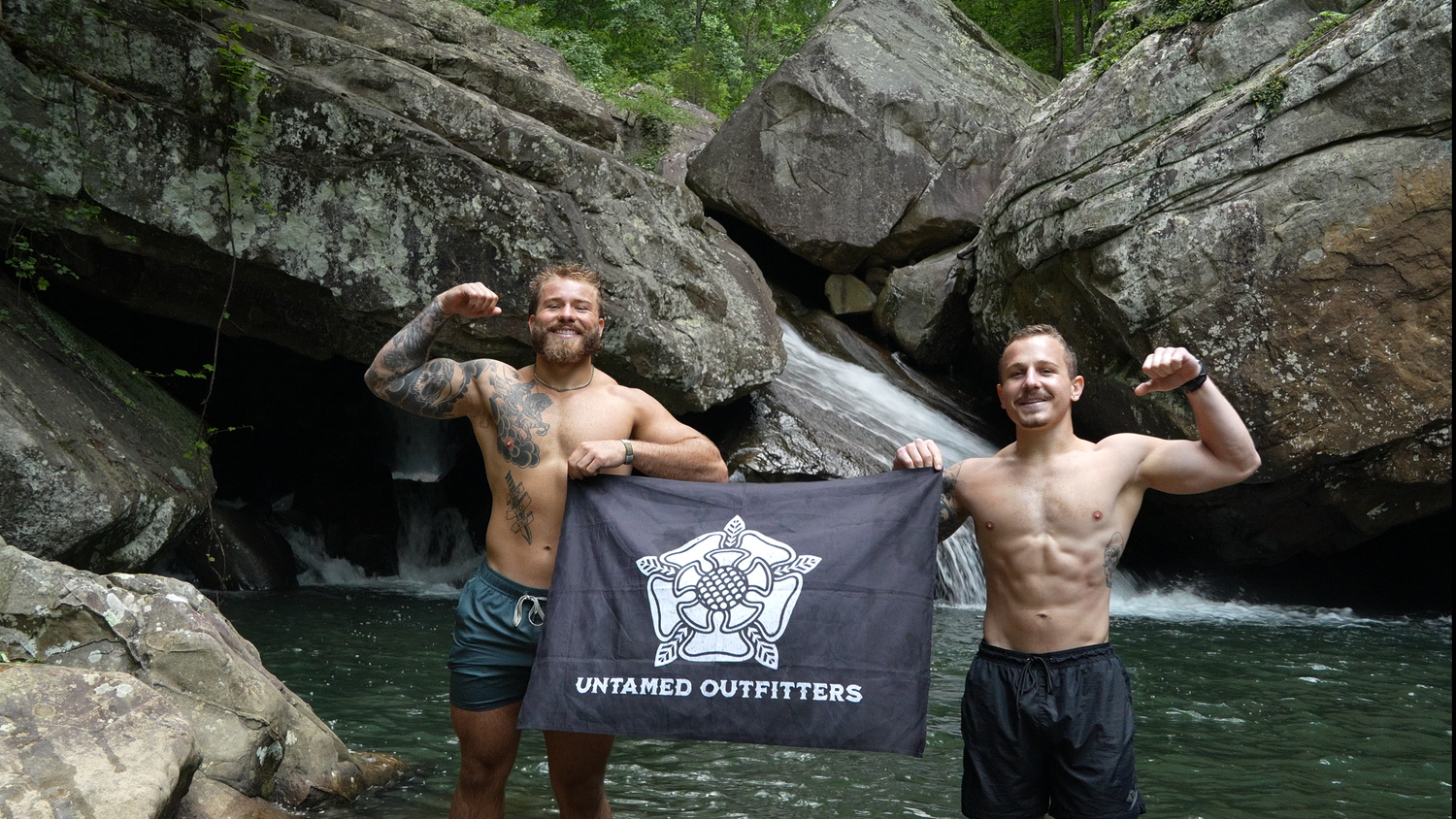 Two shirtless men flex muscles holding Untamed Outfitters flag by rocky waterfall in lush forest setting.