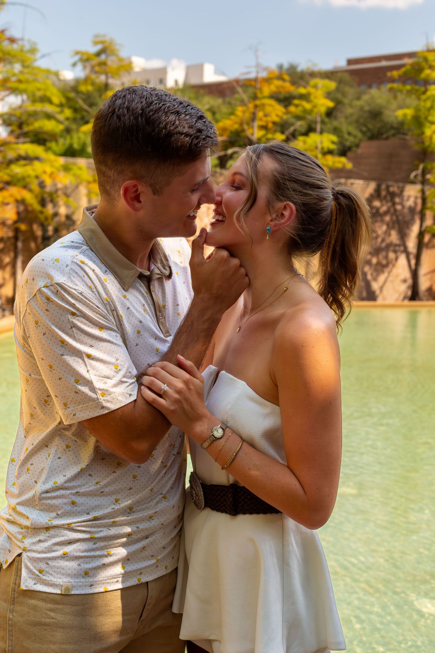 Romantic couple sharing a tender moment by a pond in a sunny park, man in casual shirt, woman in white dress.