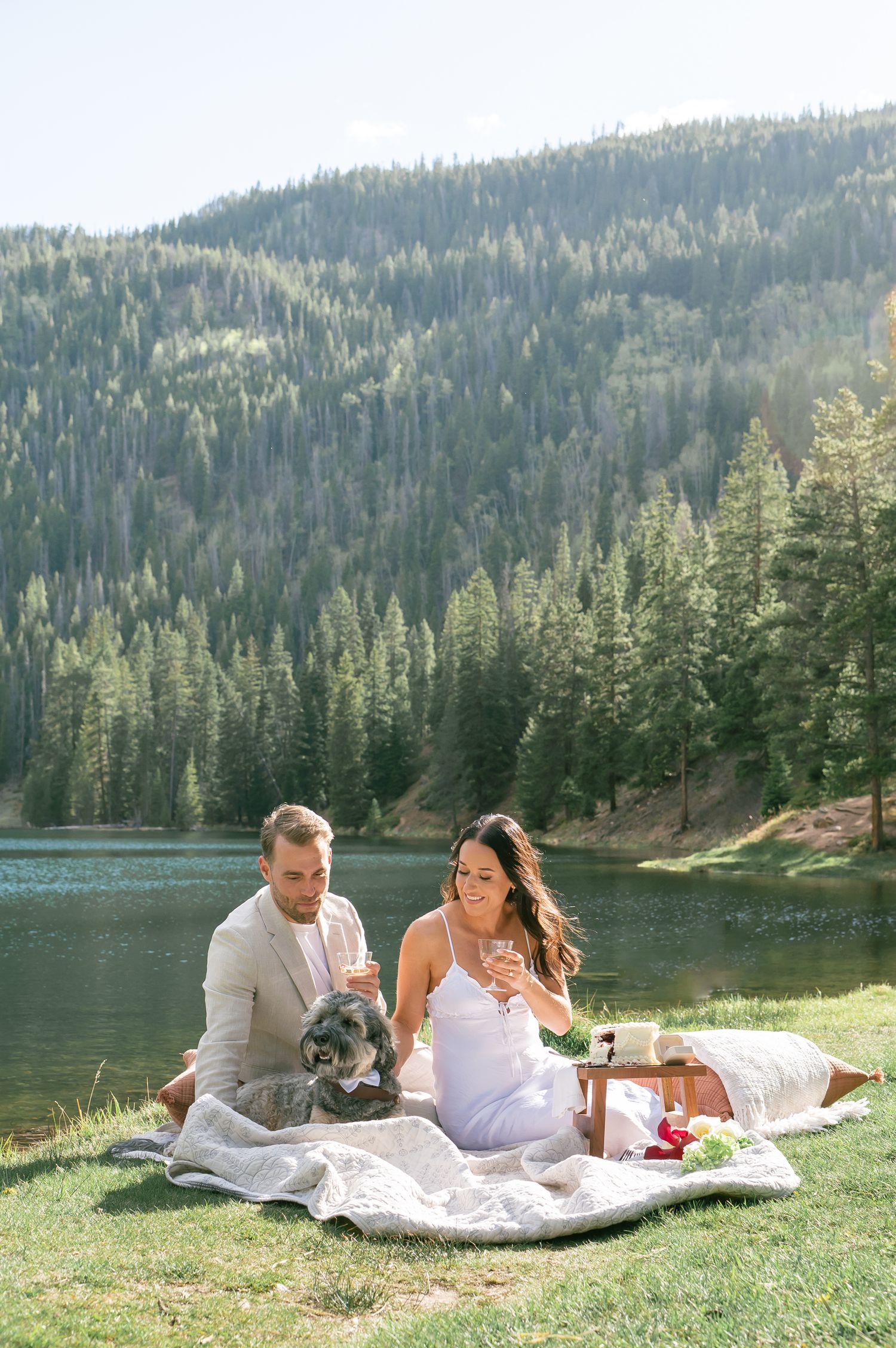 Couple enjoying a lakeside picnic with their dog, champagne, and mountain forest views on a sunny day for a Colorado elopement