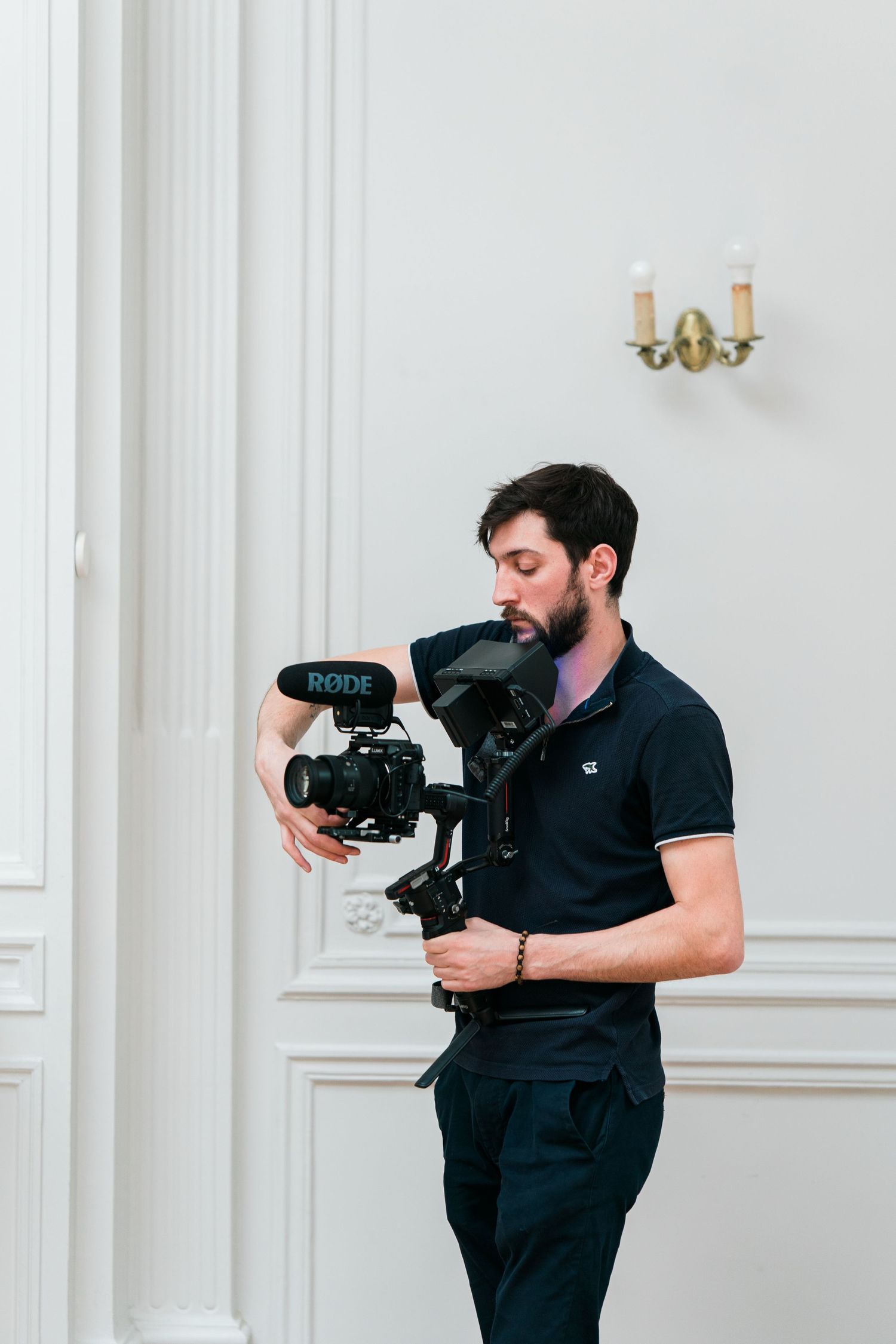 A bearded videographer holds a camera on a gimbal stabilizer with a Rode microphone in an elegant white room.
