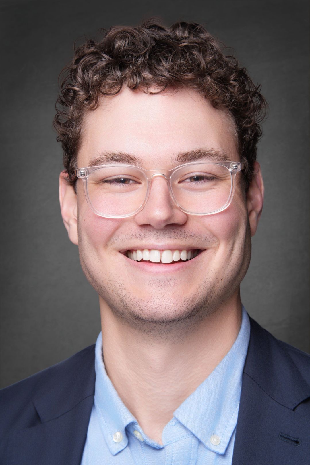 Smiling young man with curly brown hair and clear glasses wearing a navy blazer over a light blue collared shirt against a gray background.