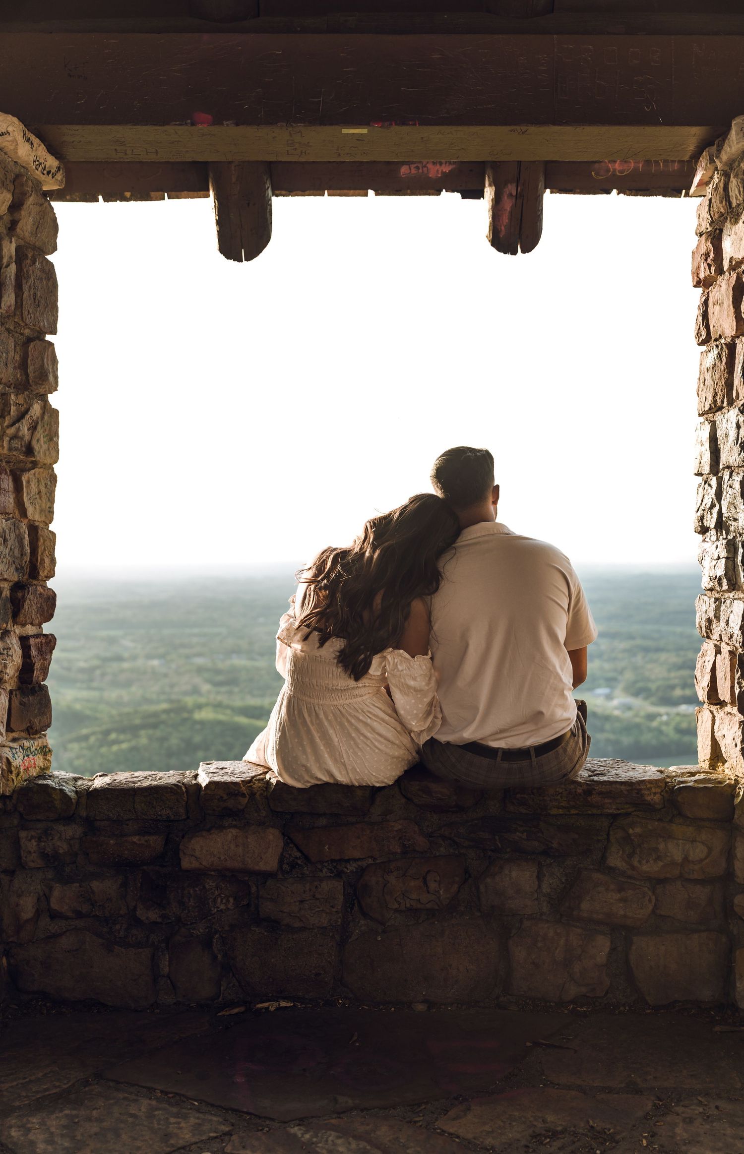 Couple sitting together on a stone ledge in a rustic tower window, gazing at a scenic green landscape view.