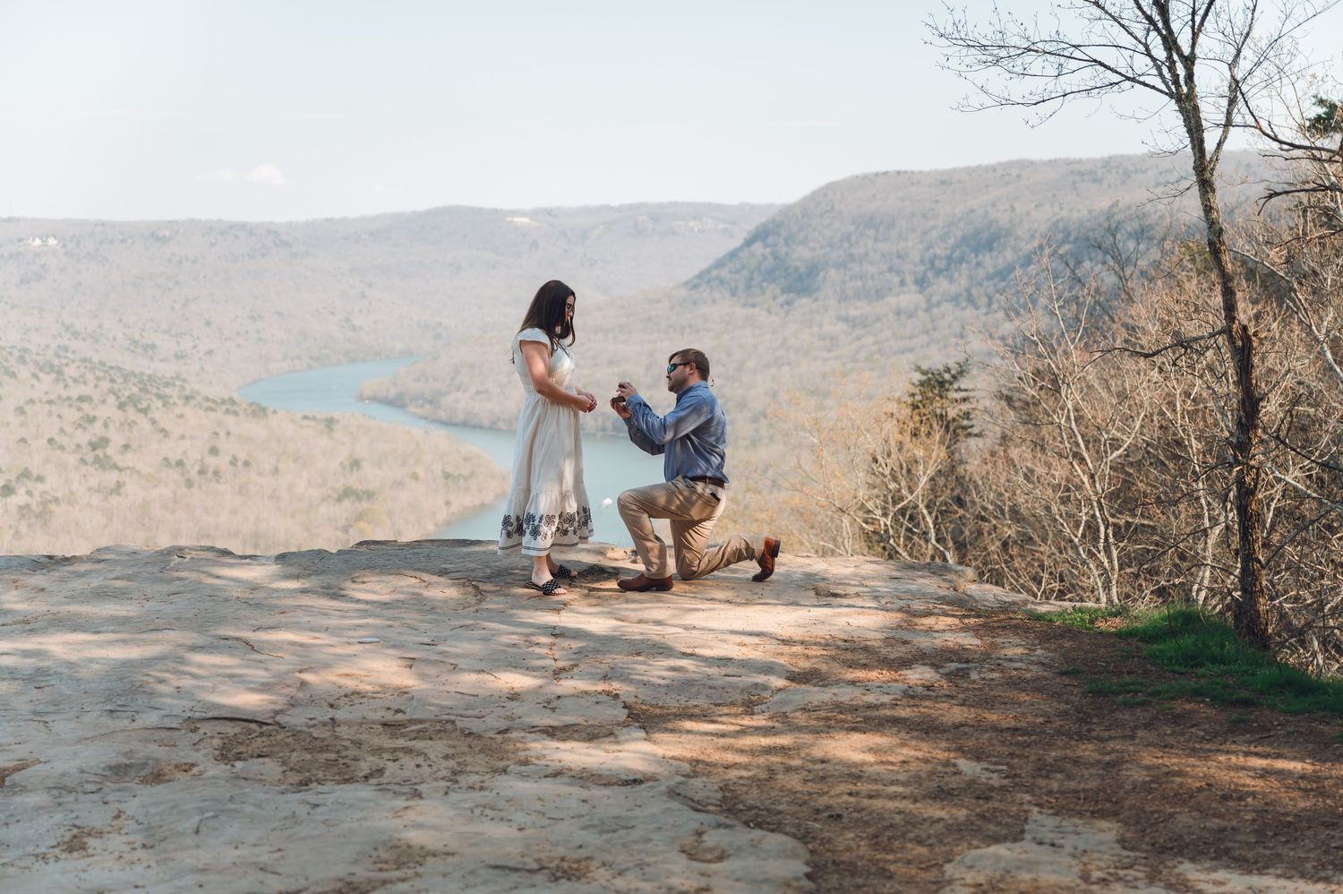Man proposes on one knee to woman on rocky overlook with scenic mountain valley and lake views.