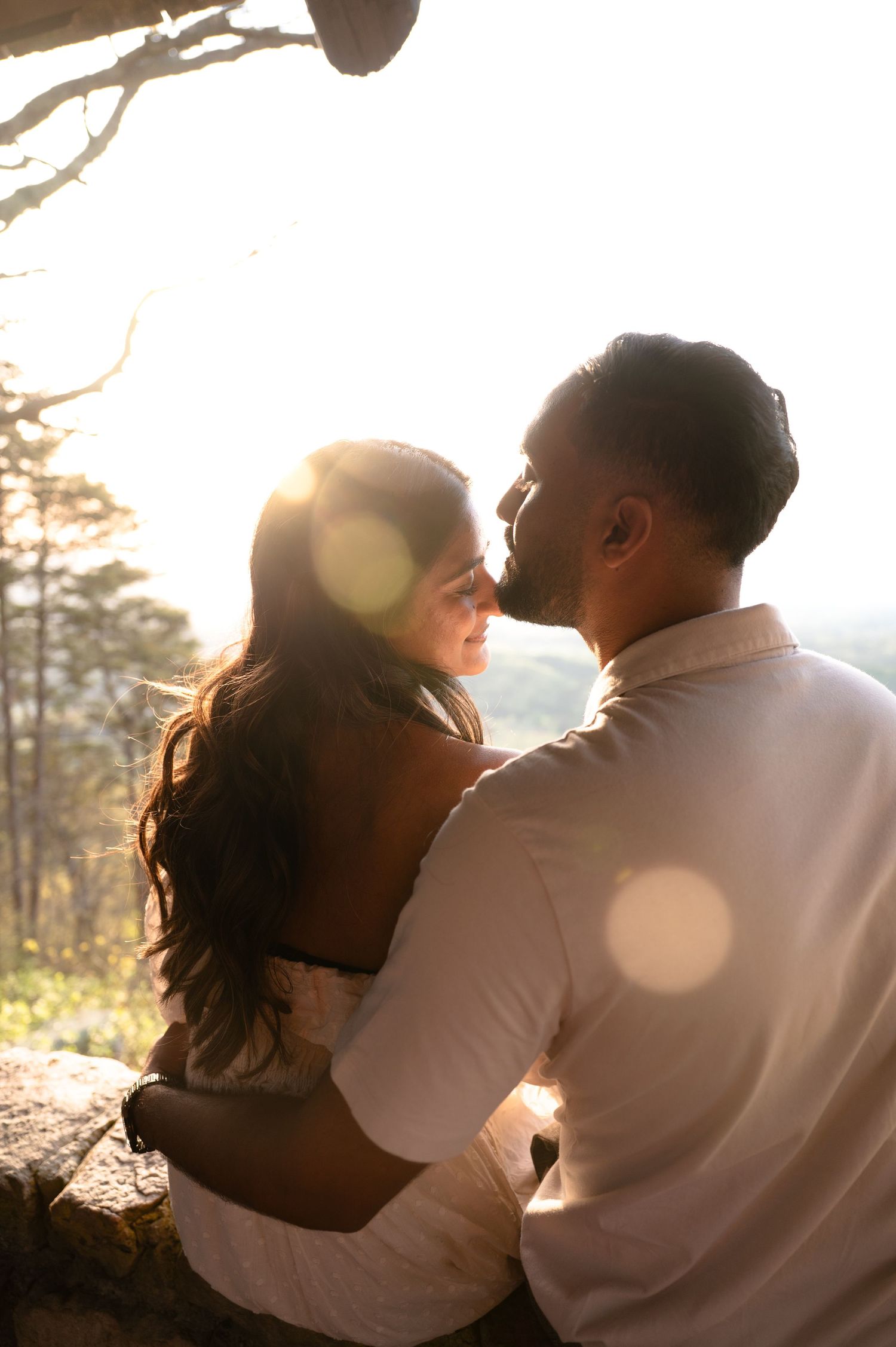 A couple embraces at golden hour, bathed in warm sunlight with a scenic forest backdrop.