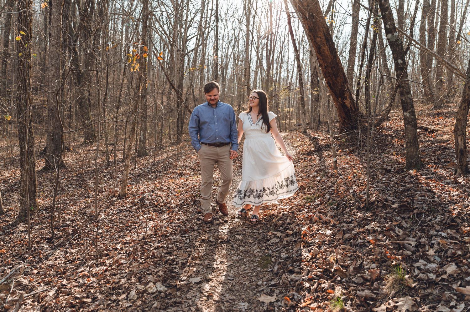 Couple walking hand-in-hand through autumn forest with fallen leaves, woman in white dress, man in blue shirt.