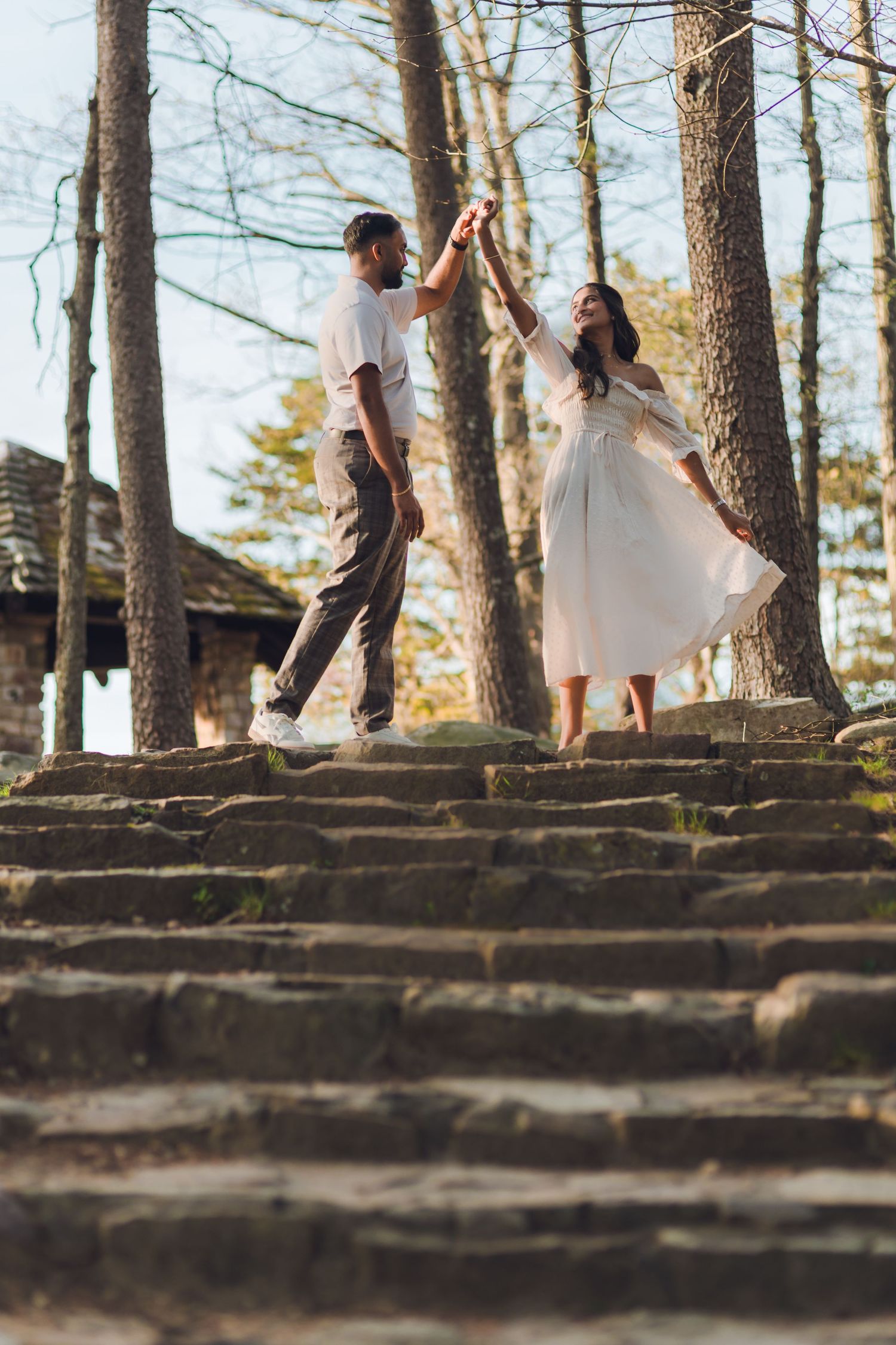 A couple holds hands on stone steps in a sunlit forest, the woman in a white dress being twirled gracefully.