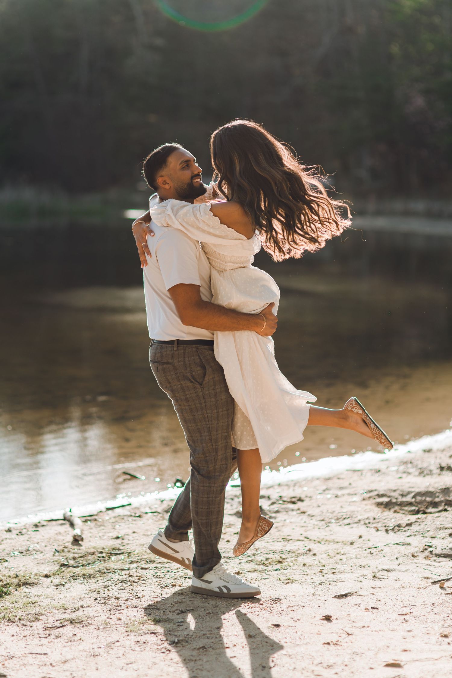 A couple embraces joyfully by a sunlit riverside, the man lifting the woman in a white dress as her hair flows.
