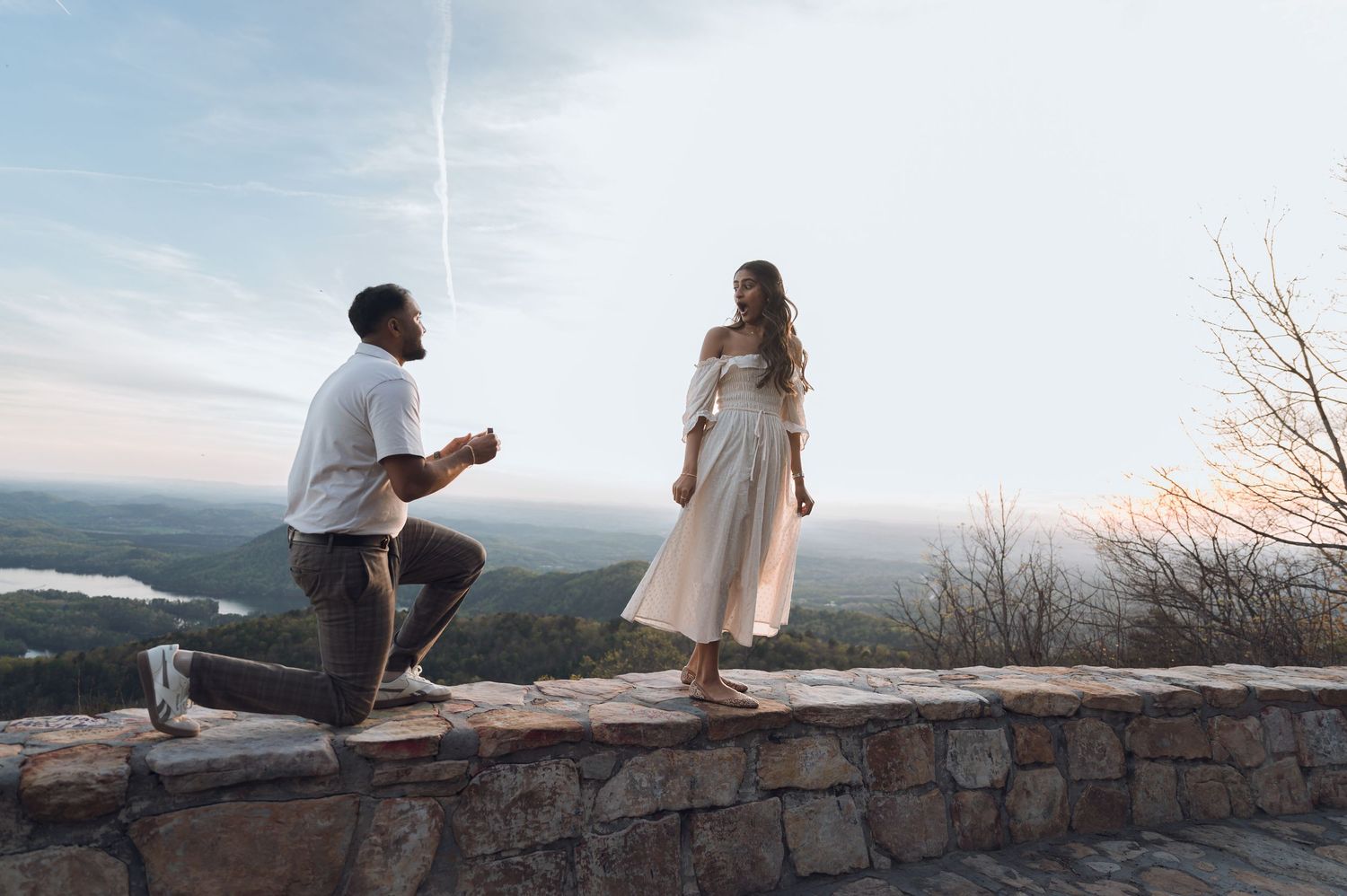 Man kneeling on stone wall proposing to woman in white dress at scenic overlook with panoramic landscape views.