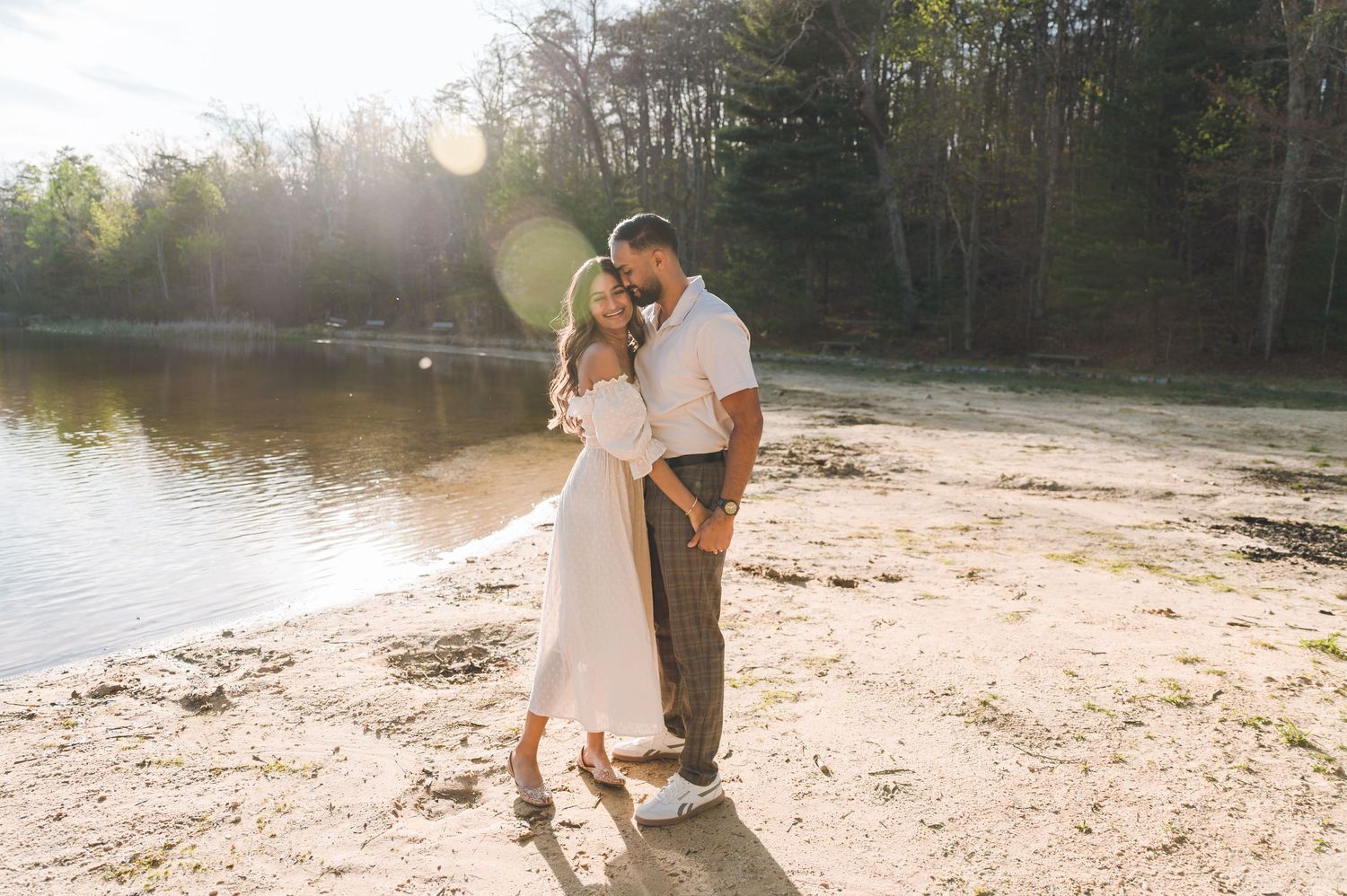 Couple embracing romantically by a sunlit lakeside beach surrounded by lush green trees.