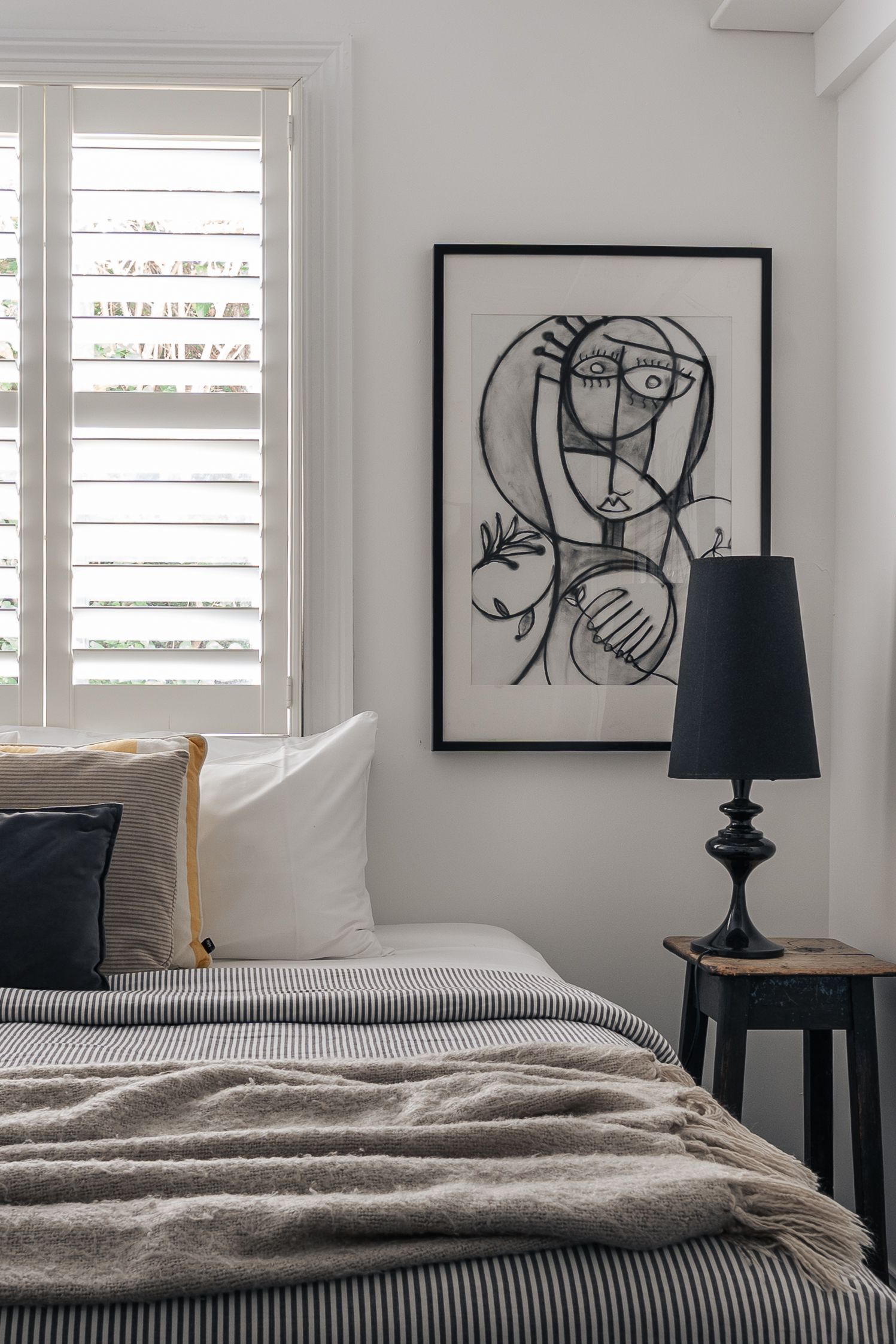 Minimalist bedroom with striped bedding, black framed line art portrait, white shutters, and black table lamp.

Piccadilly House