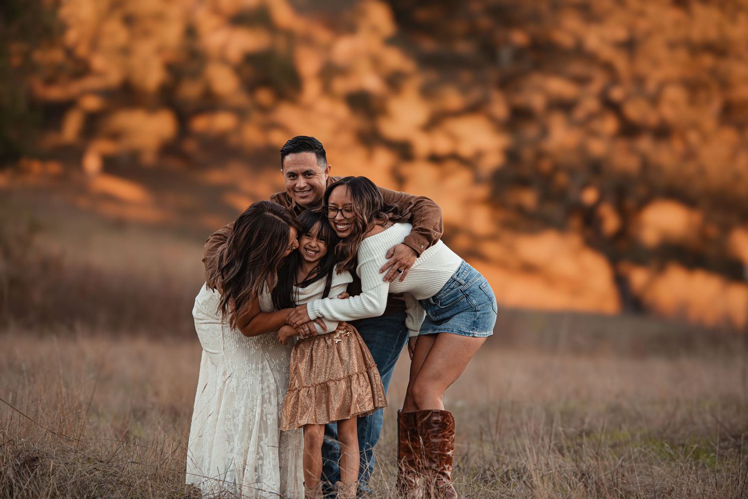 Happy family of four sharing a warm group hug in a golden sunlit field during an outdoor photo session.