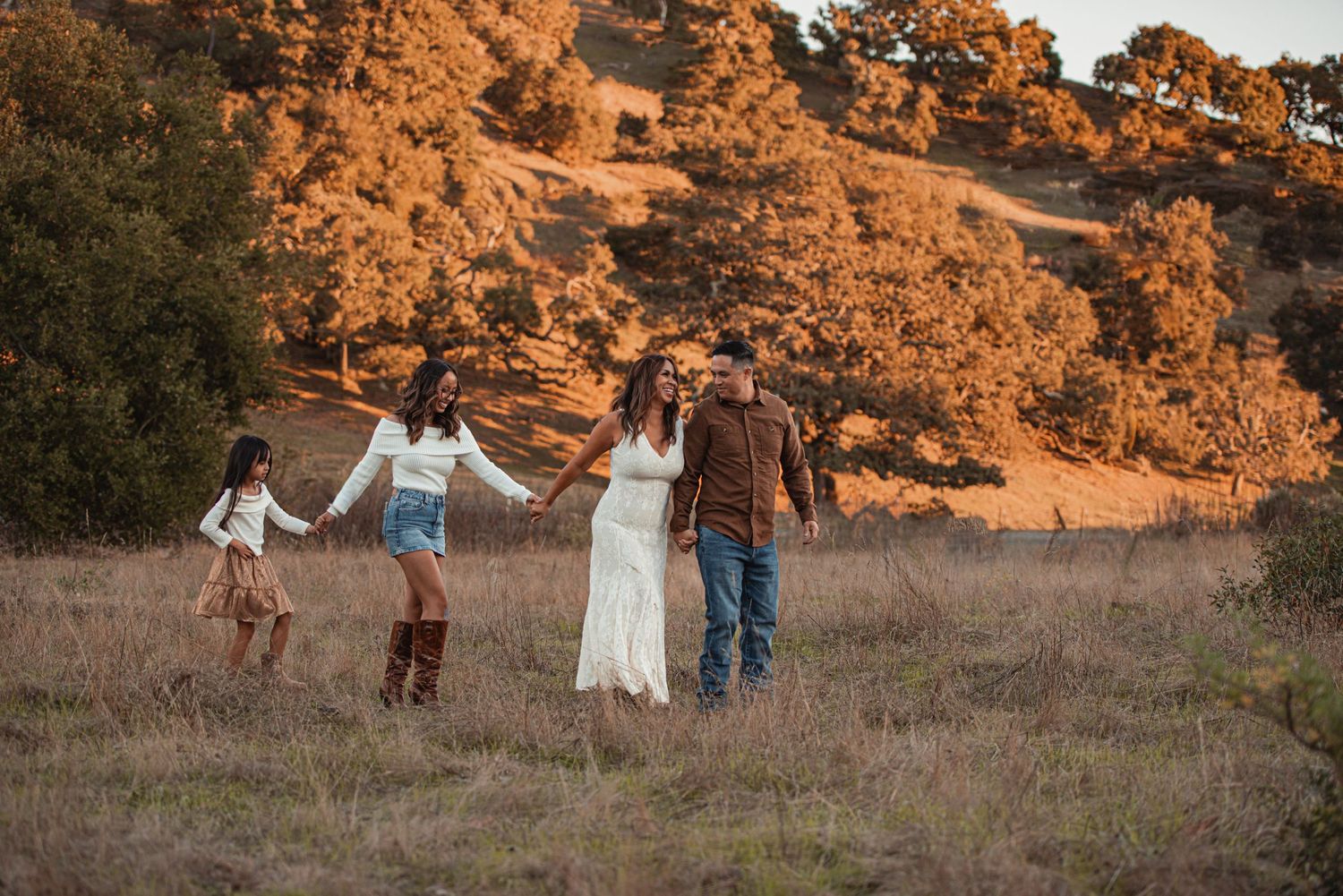 Family of four walking through golden fields at sunset with autumn trees in the background.
