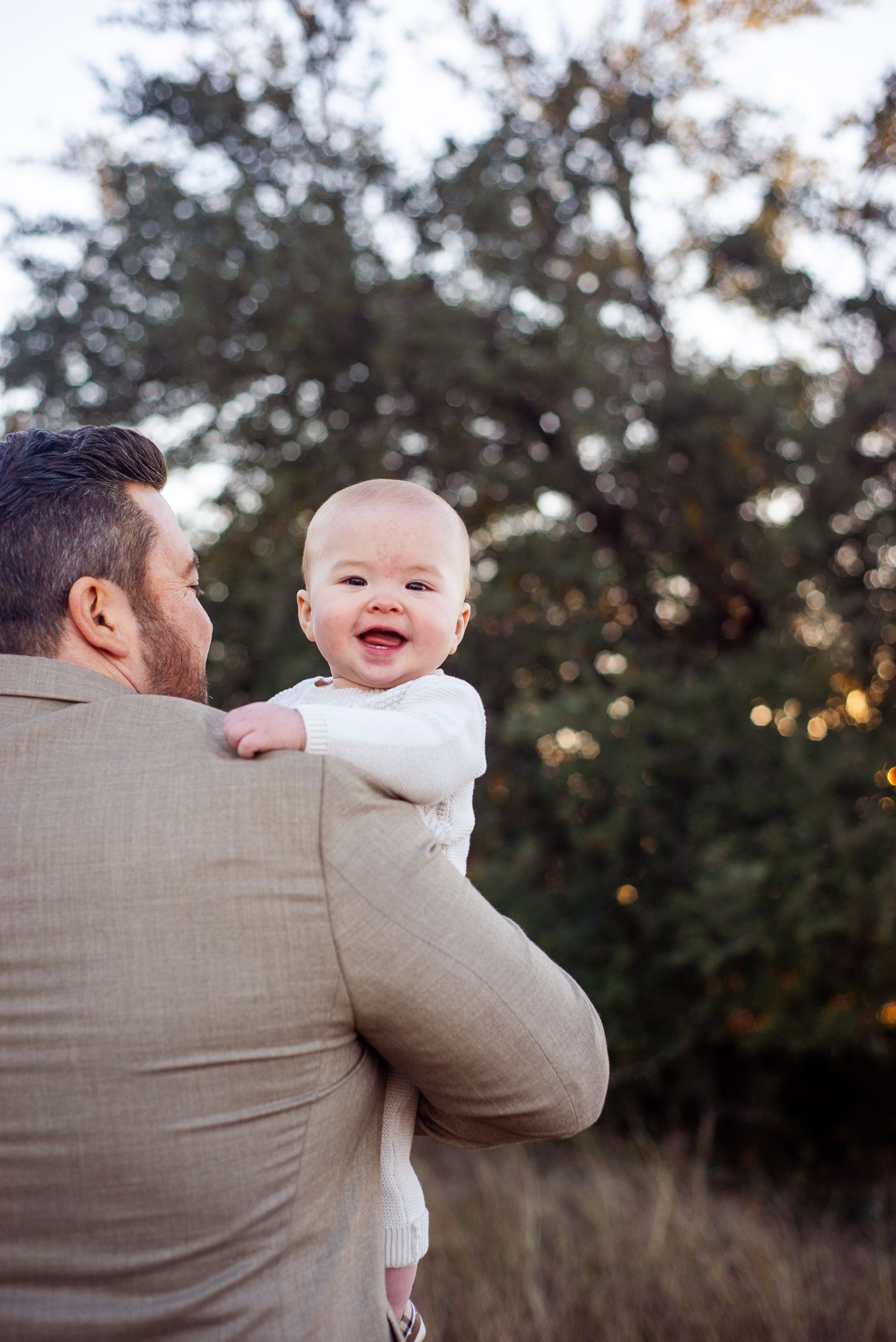 Baby in white outfit smiling while being held outdoors at sunset.