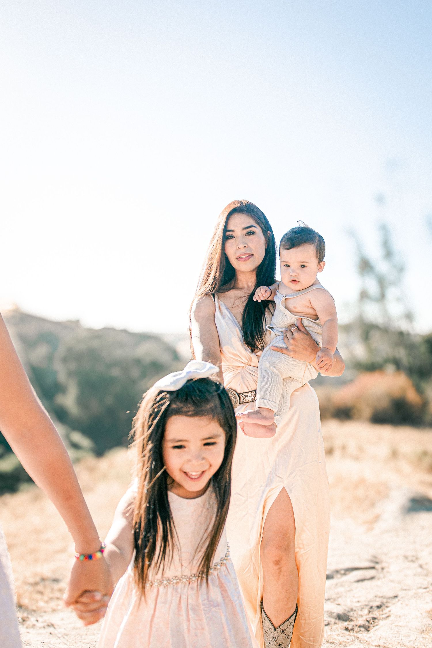 A woman in a flowy dress holds a baby while a young girl smiles outdoors in a sunny, arid landscape.