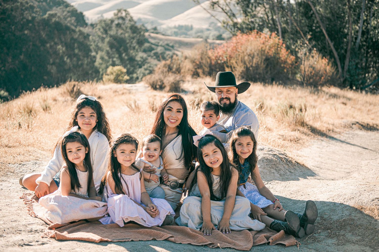 Large Hispanic family posing outdoors on a blanket in a golden hillside landscape during a sunny day.