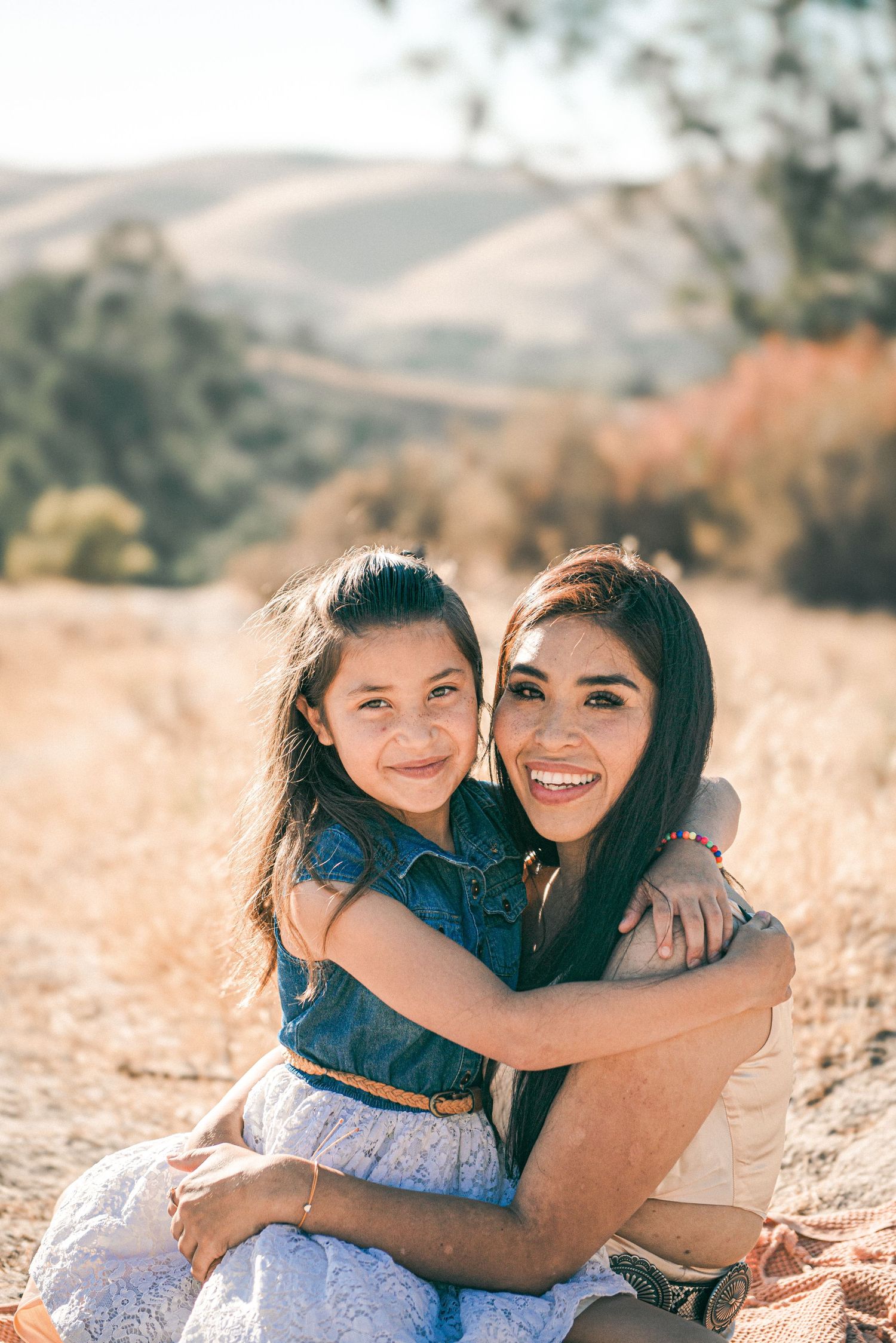 A smiling woman and young girl hug outdoors in a sunny field with golden grass and rolling hills in the background.