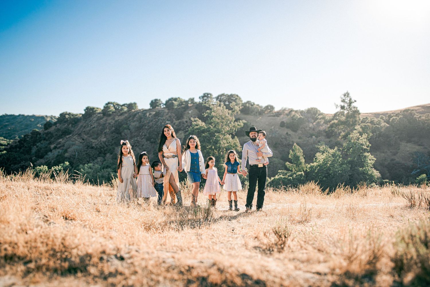 Large family group posing in a sunny golden field with green hills and clear blue sky in the background.