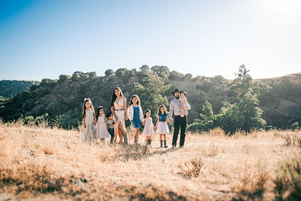 Large family group posing in a sunny golden field with green hills and clear blue sky in the background.