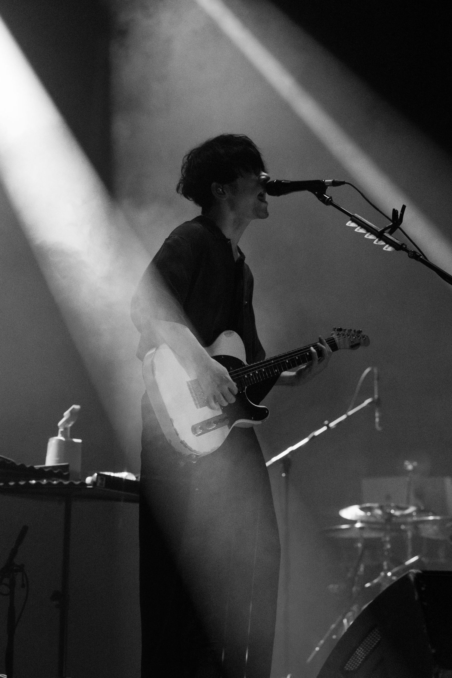 Black and White image of TK from Ling Tosite Sigure singing into a mic on stage at the Belasco with 2 beams of concert lights crossing him.