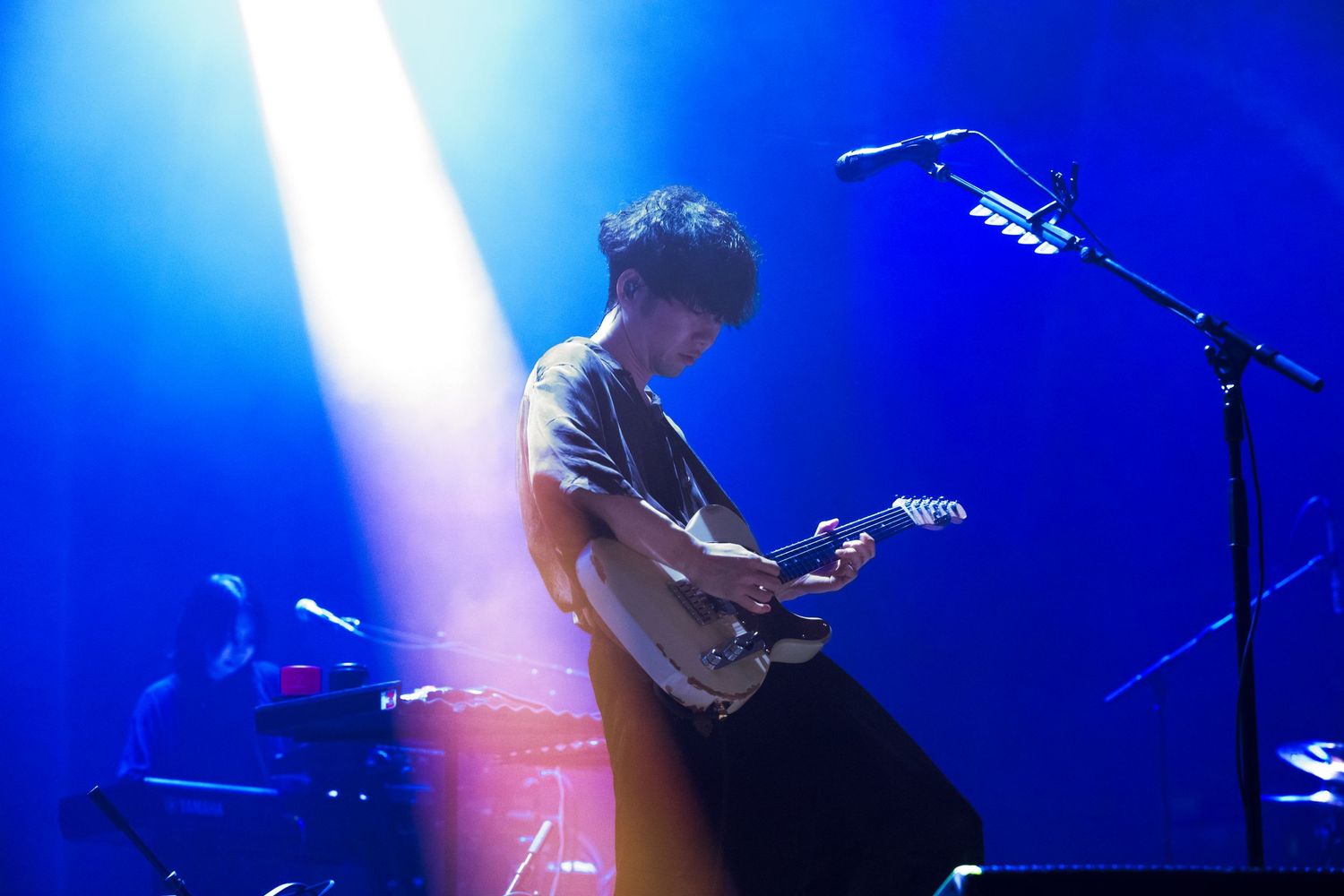A photo of TK from Ling Tosite Sigure underneath blue concert lights during his performance at The Belasco. He is playing a guitar on stage.