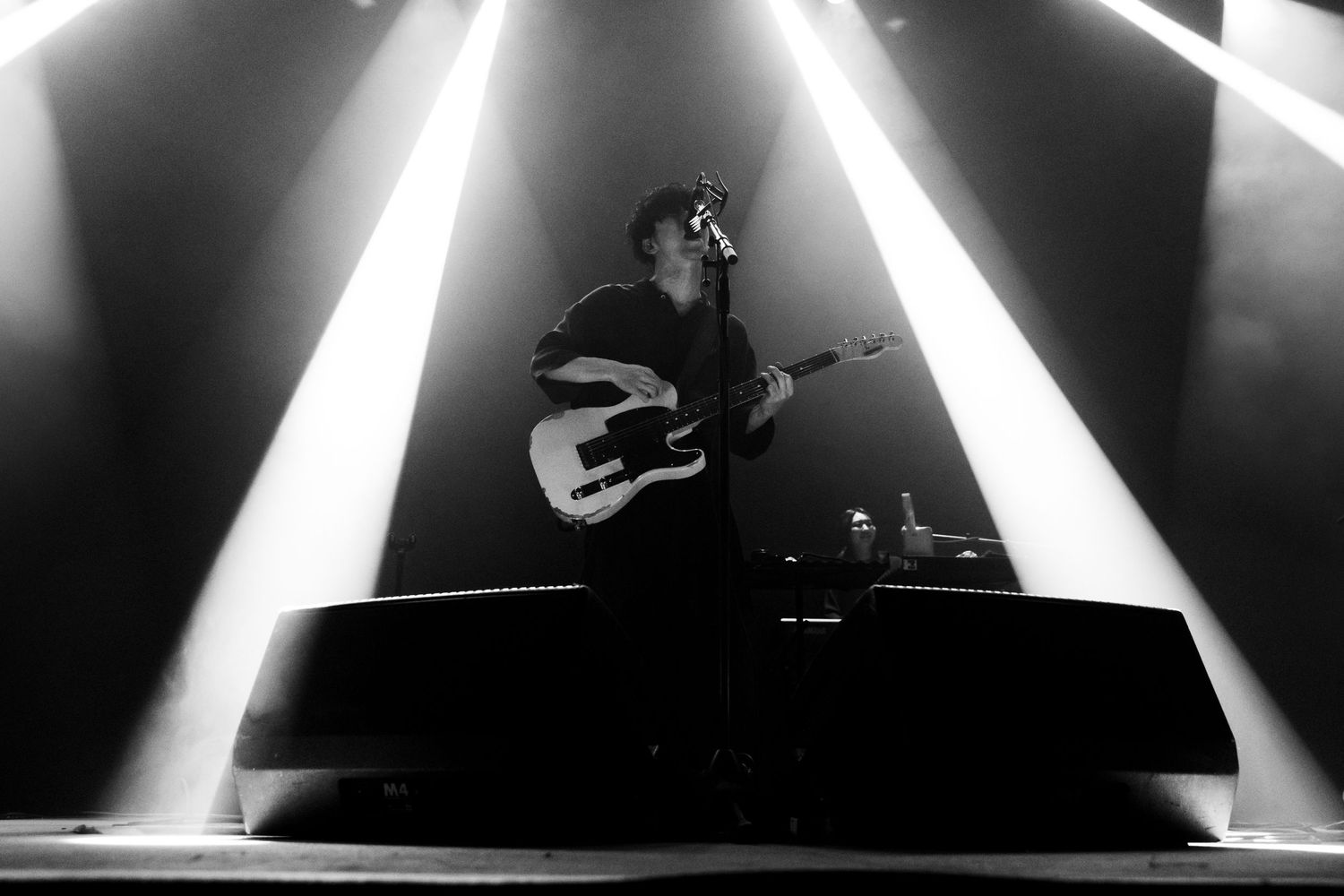 A black and white photo of TK from Ling Tosite Sigure concert lights during his performance at The Belasco. He is playing a guitar on stage.