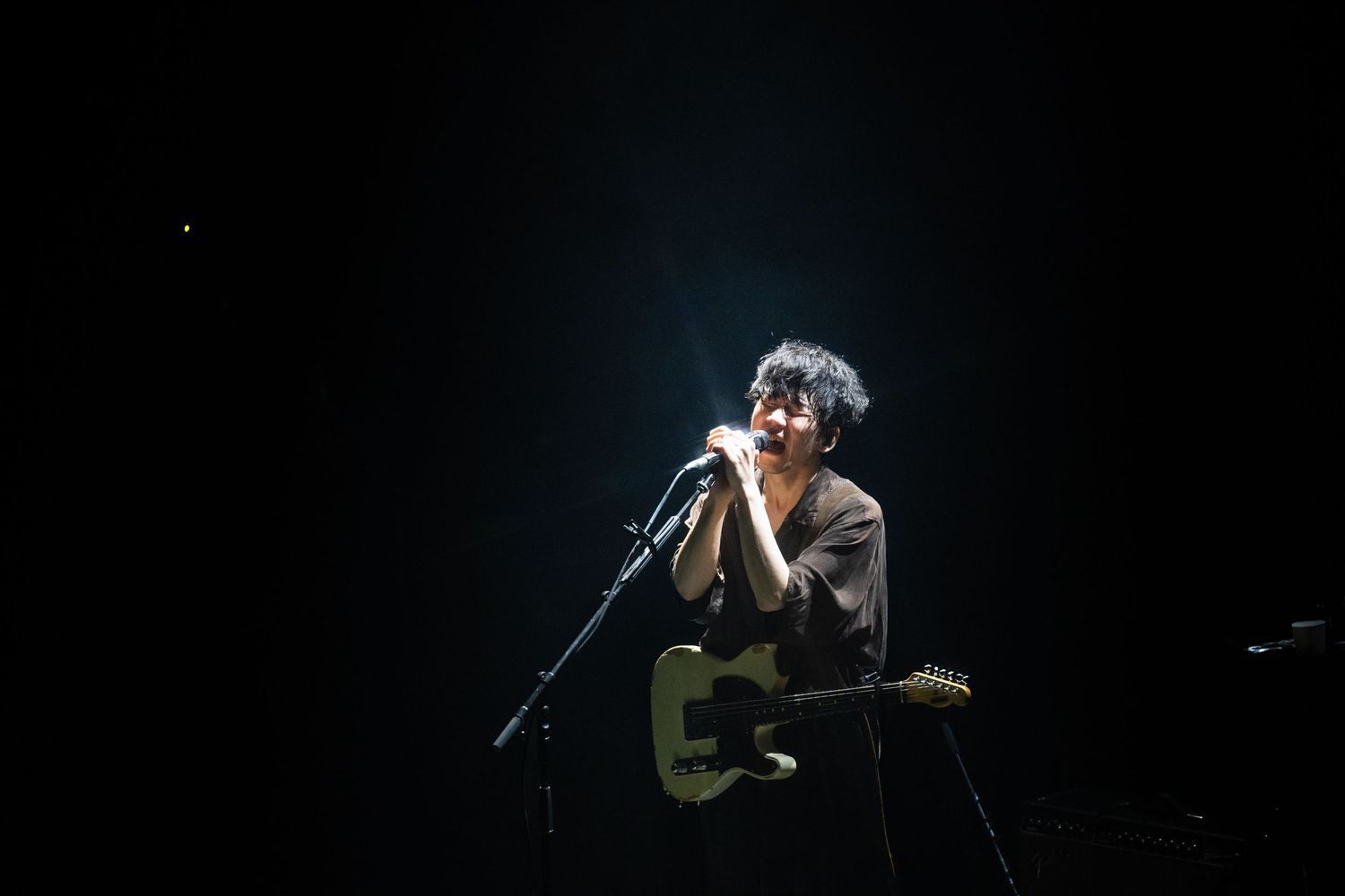 TK from Ling Tosite Sigure performing a concert live at the Belasco. Tk sings into a mic on stage surrounded by the dark.