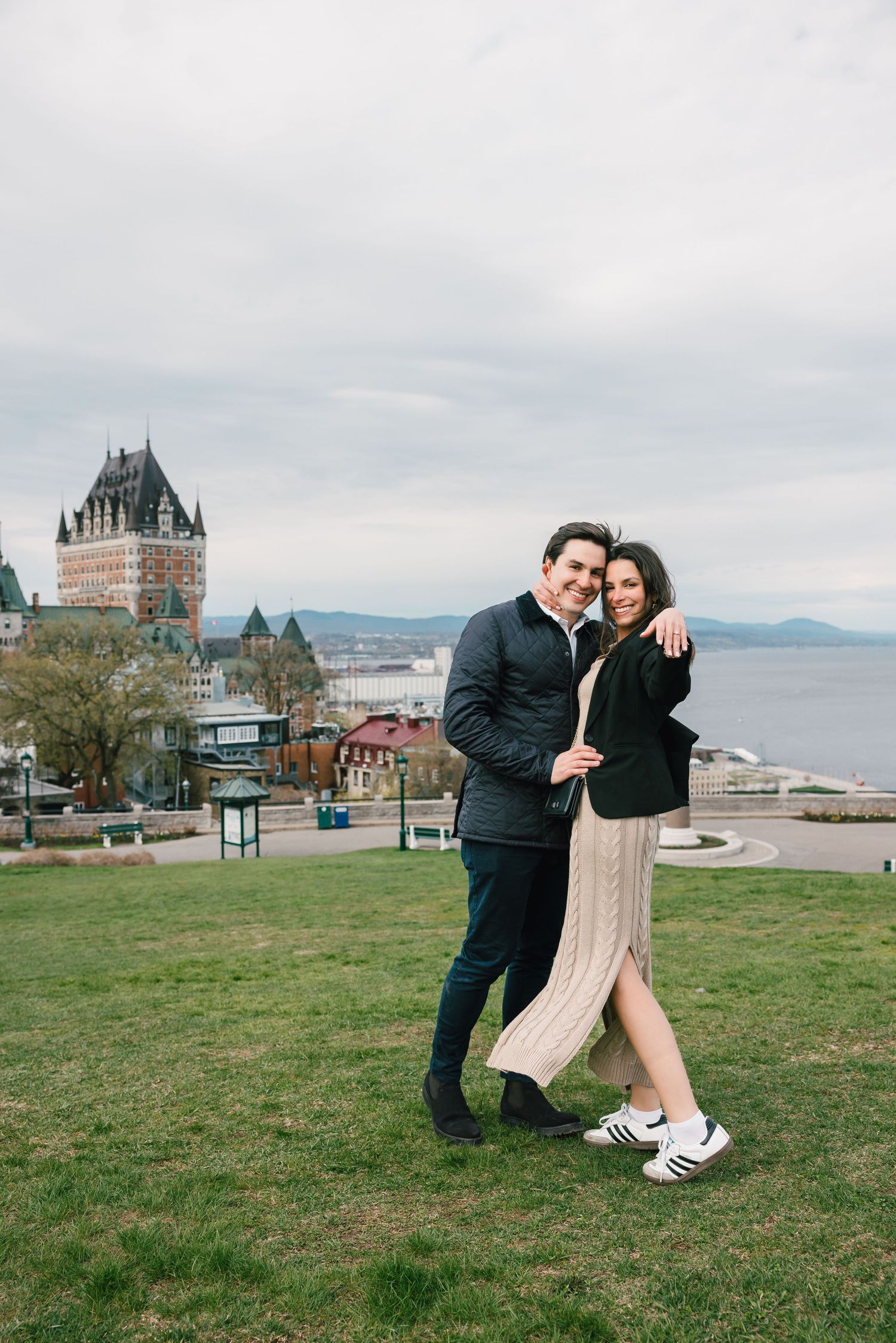 A couple walks hand in hand on a grassy hill overlooking Chateau Frontenac and the historic Quebec City waterfront.