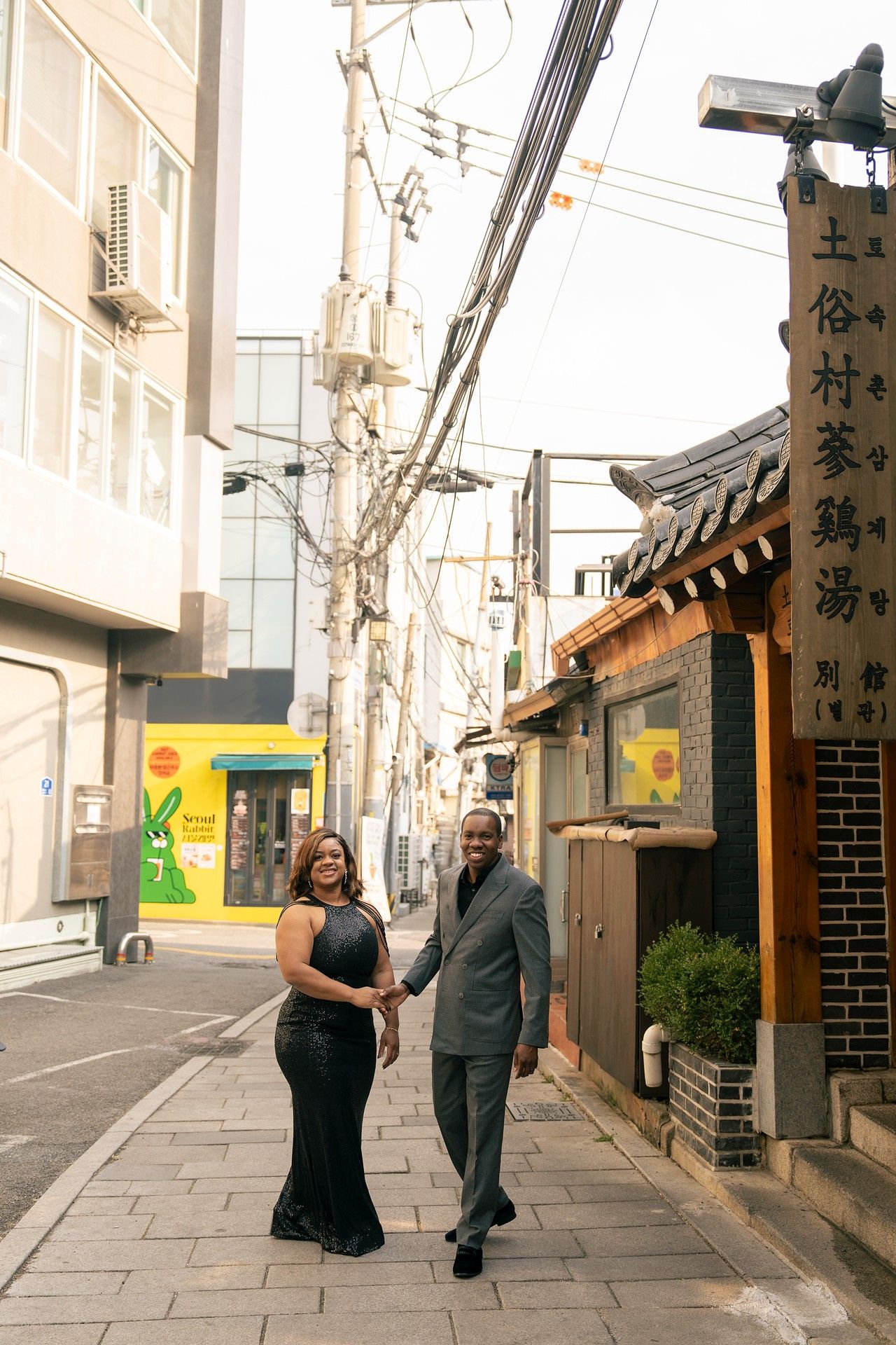 Couple holding hands and smiling in a Korean alley, woman in black gown and man in gray suit, traditional signage nearby.