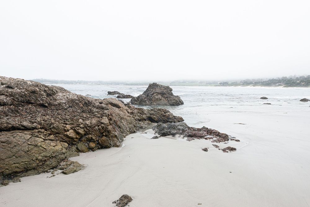 Rocky shoreline with sandy beach on a misty overcast day, with calm ocean waters and distant foggy hills.