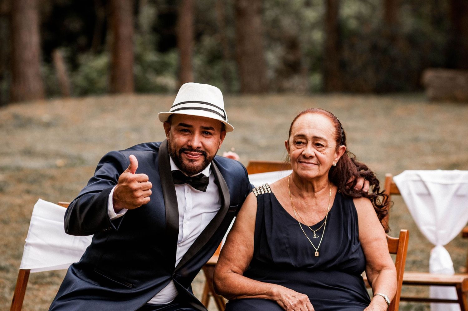 Smiling groom in tuxedo giving thumbs up seated beside elderly woman at outdoor woodland wedding ceremony.
