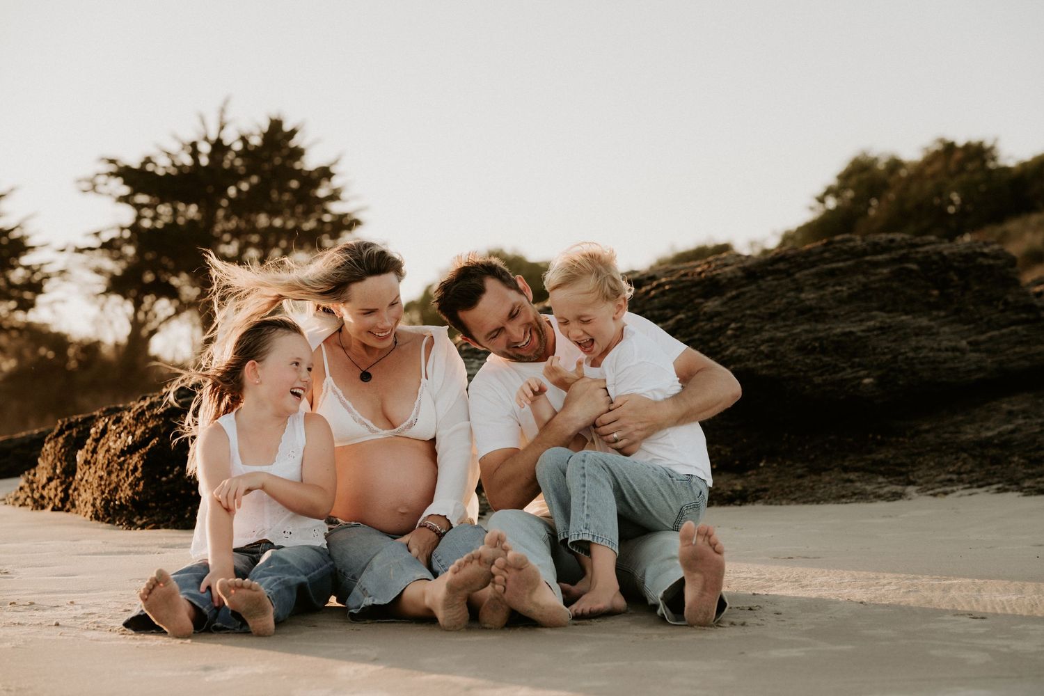 Happy family of four sitting together on a sandy beach at golden hour, laughing and embracing warmly.