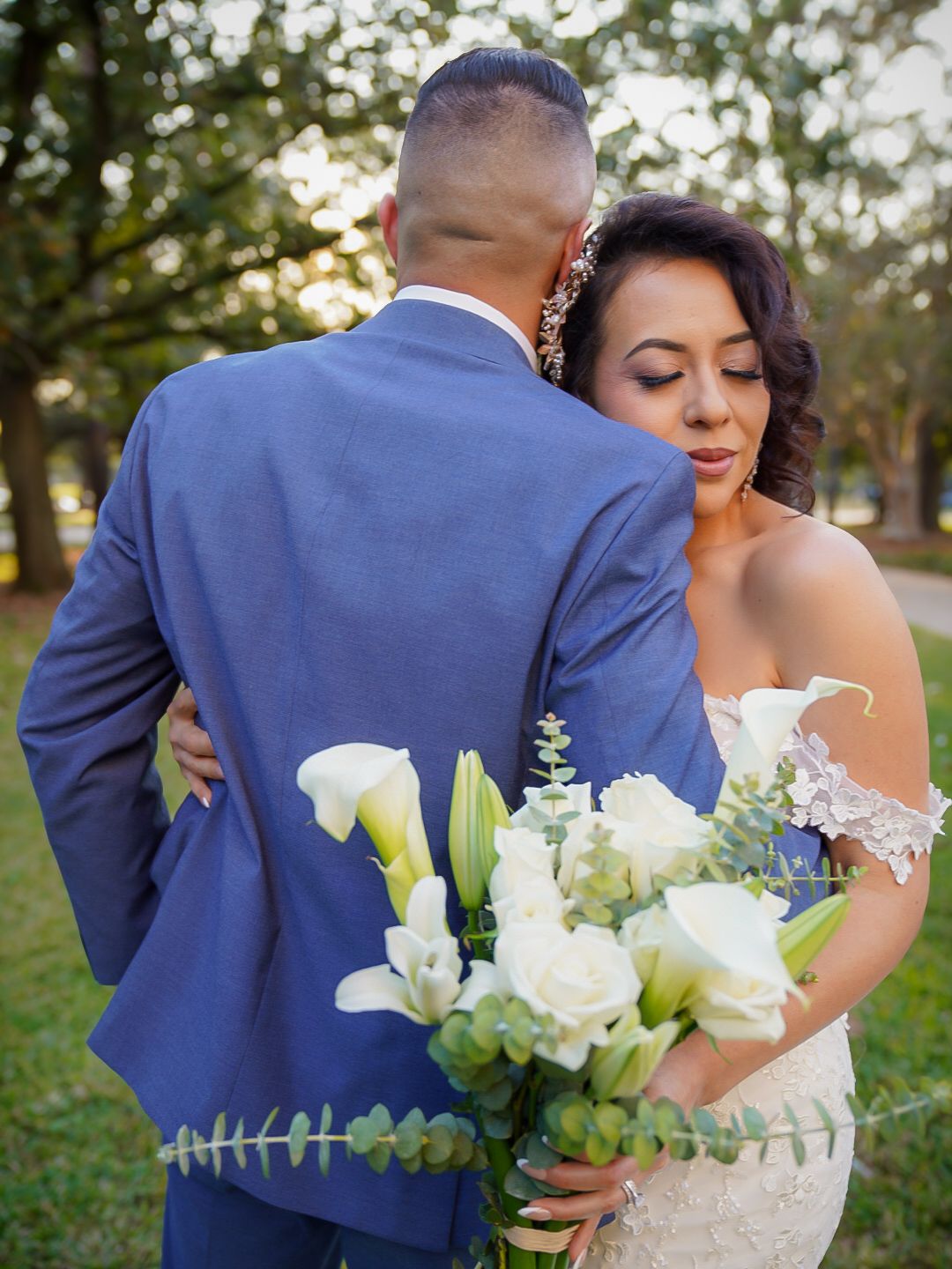 Bride and groom share romantic moment, she holds white floral bouquet, he wears blue suit, evening outdoor wedding photo.