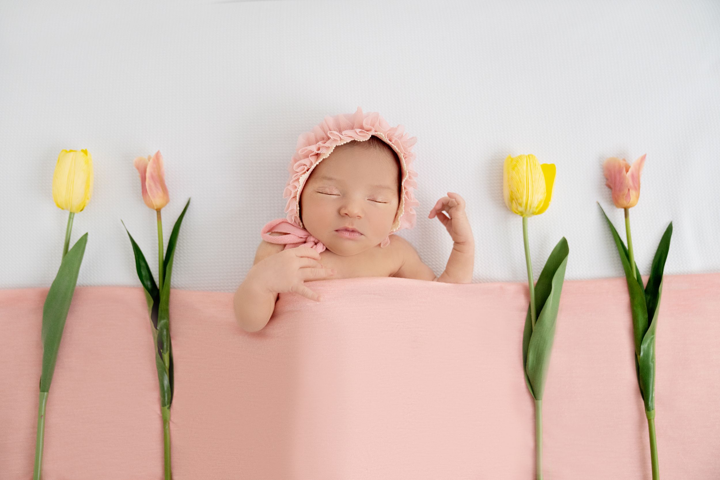 Sleeping newborn baby in pink bonnet surrounded by yellow and pink tulips on a soft pink and white background.