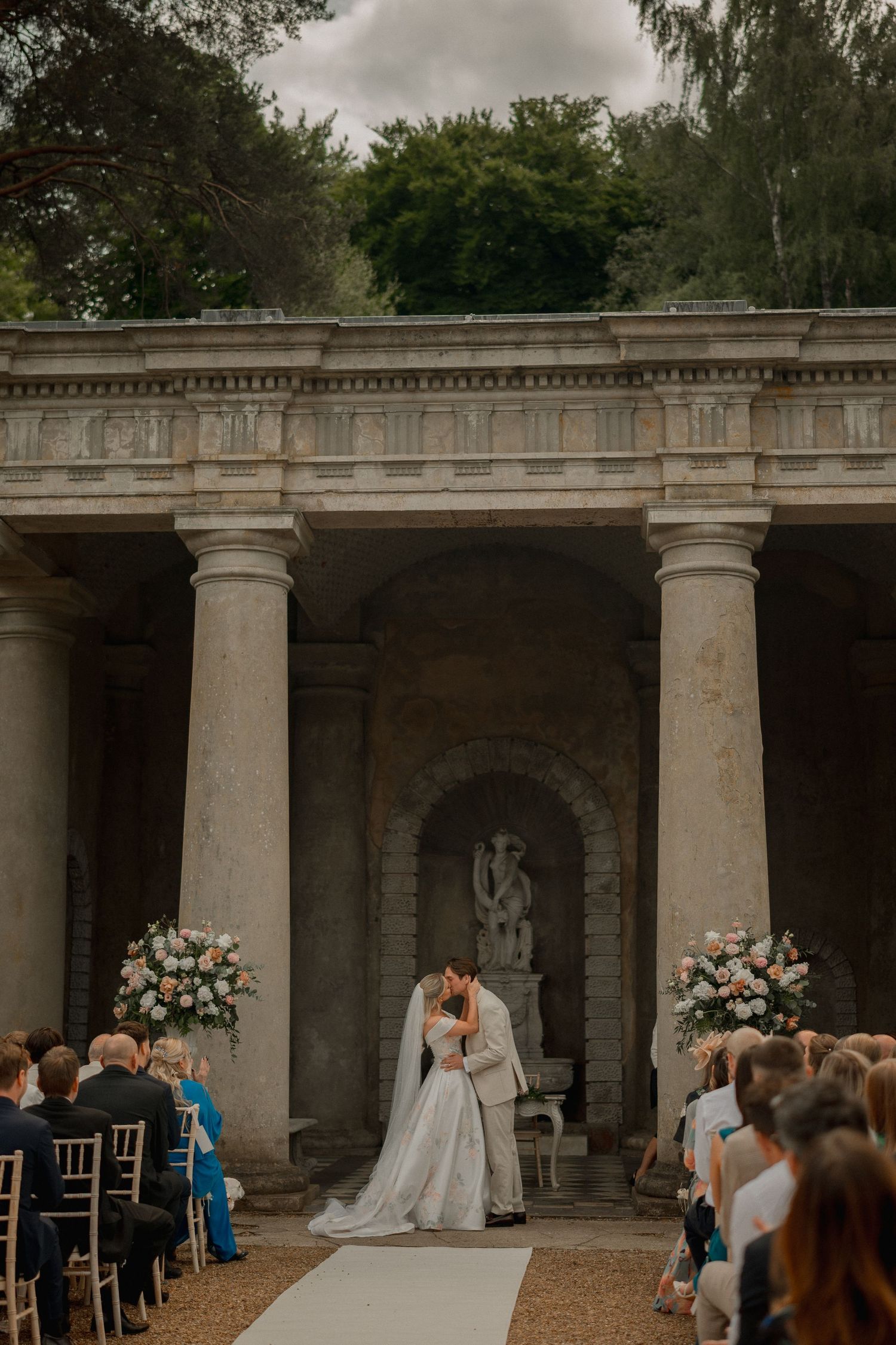 Bride and groom share first kiss at outdoor wedding ceremony beneath classical stone colonnade with floral arrangements.