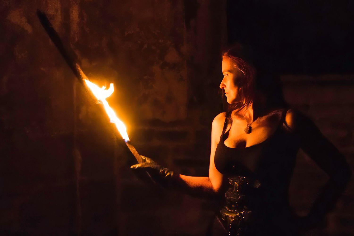 A woman in a black corset holds a flaming torch in a dark, dramatic setting, illuminated by the fire's warm glow.