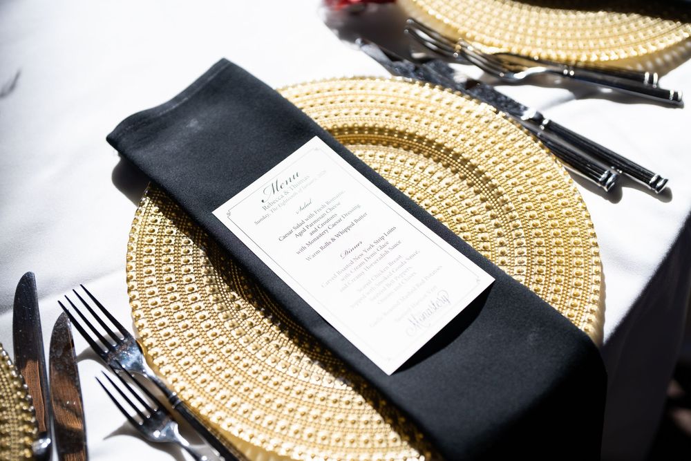 Elegant gold charger plate with black napkin and menu card, formal table setting with black silverware on white tablecloth.