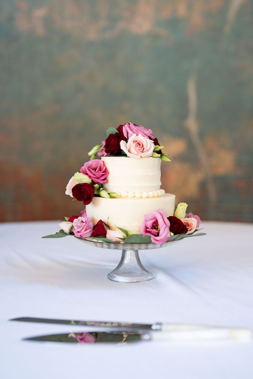 Two-tier white wedding cake with pearl trim, adorned with pink and burgundy roses on a silver cake stand.