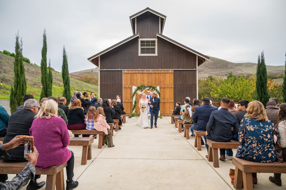 Outdoor wedding ceremony at a rustic barn venue with guests seated on wooden pews as couple walks down the aisle.