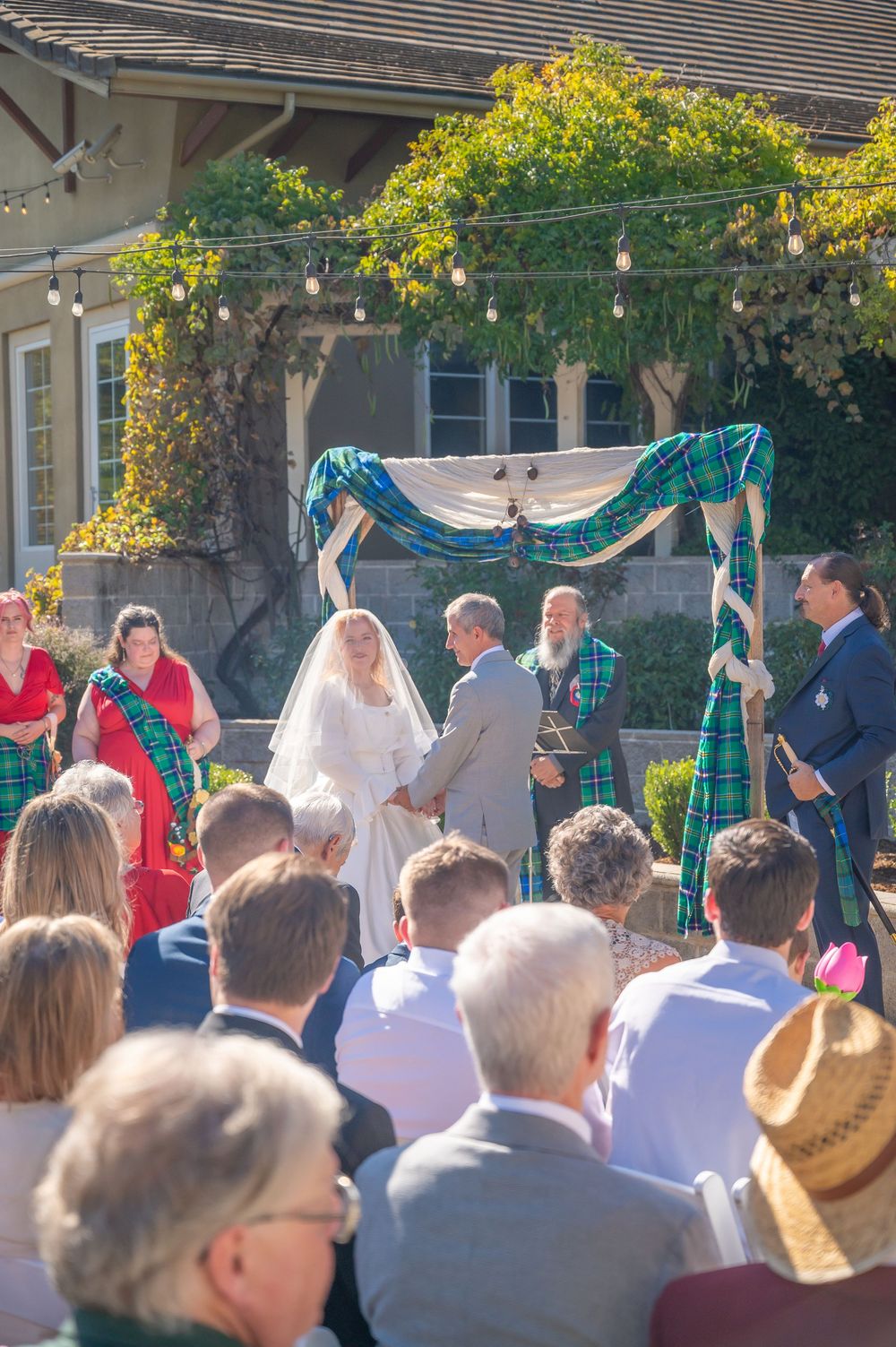 Outdoor wedding ceremony with bride and groom under a tartan-draped arch, surrounded by seated guests in a garden setting.
