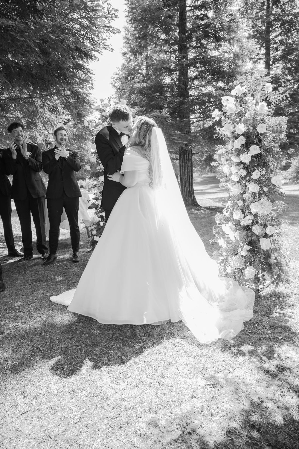 Black and white photo of a bride and groom sharing their first kiss at an outdoor wedding ceremony among tall trees.