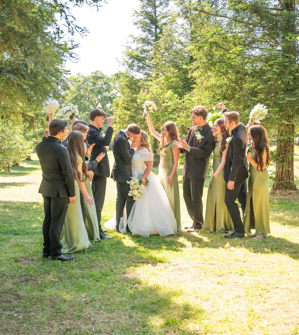 Wedding party in green dresses and black suits surrounds kissing bride and groom outdoors in sunny garden.